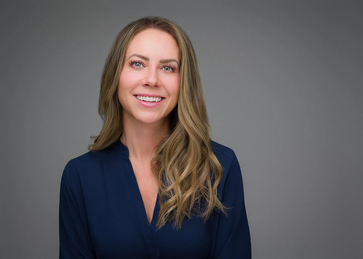 A woman with long wavy light brown hair and blue eyes smiling, wearing a dark blue top, against a gray background.