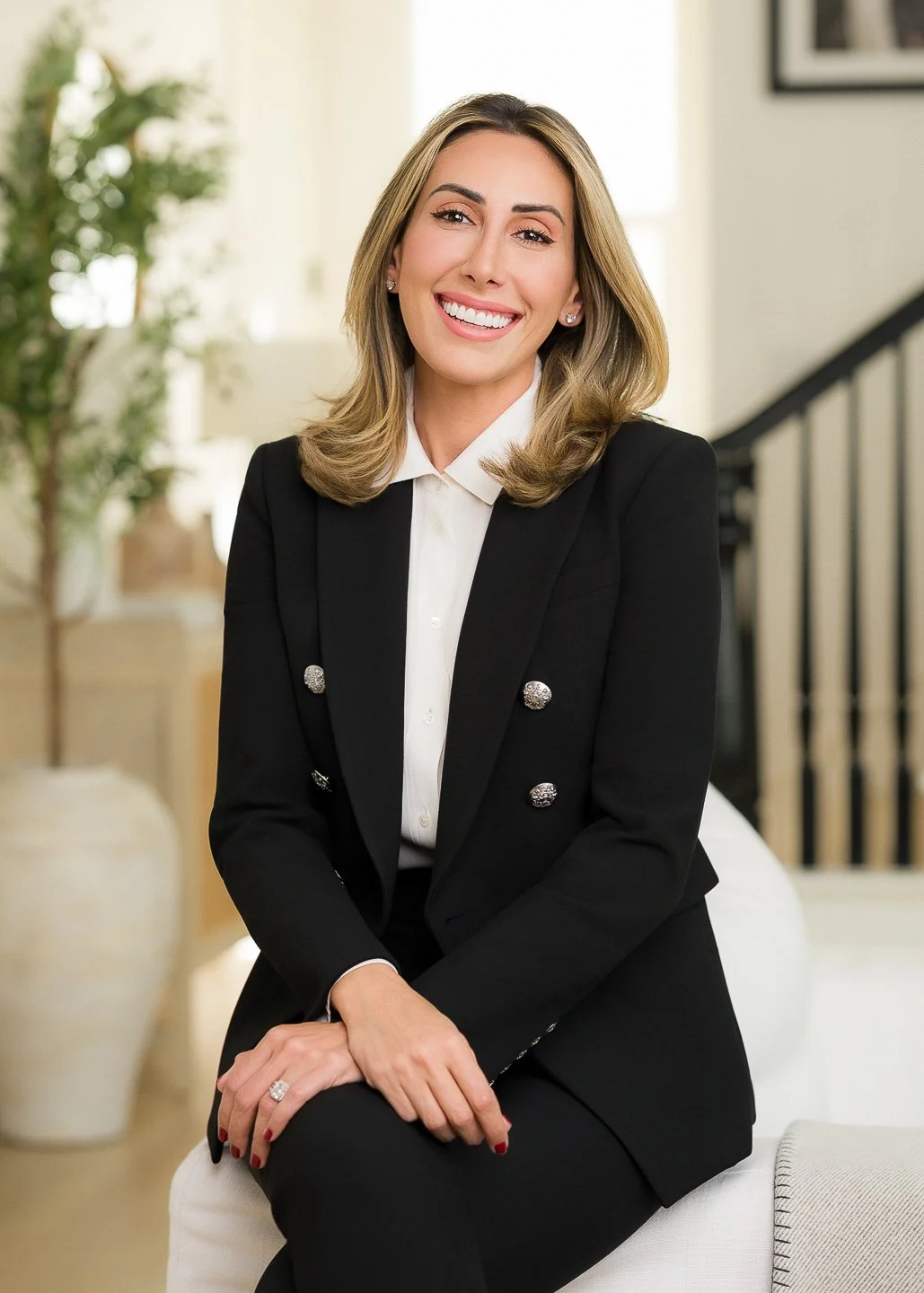 A smiling woman with shoulder-length blonde hair, wearing a black blazer with silver buttons and a white blouse, sitting indoors on a white chair.