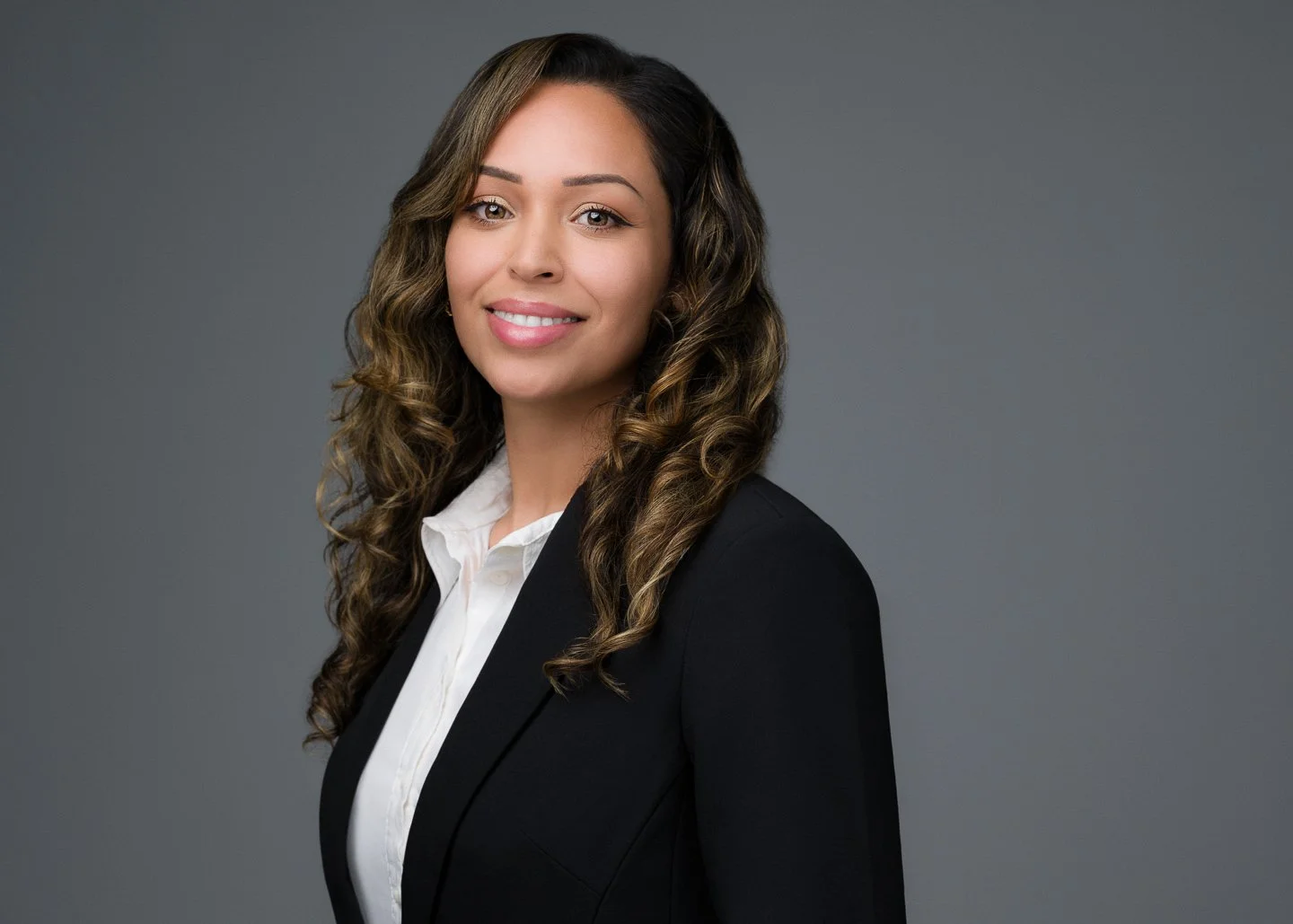 A professional woman with long, curly hair smiling at the camera, dressed in a black blazer and white blouse against a gray background.