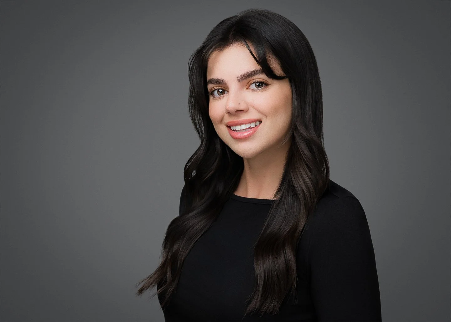 A young woman with long dark wavy hair, smiling, wearing a black top, standing against a gray background.