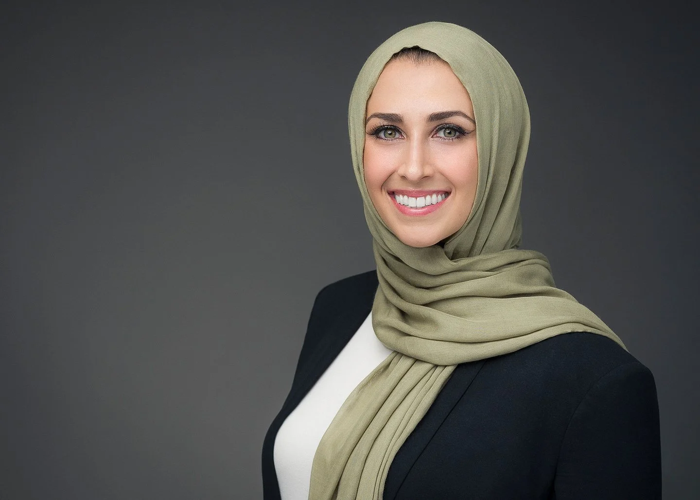 A woman wearing a light green hijab, a black blazer, and a white top, smiling against a gray background.