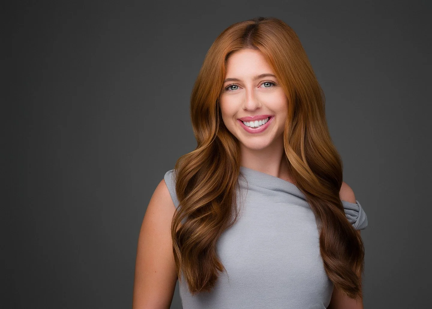 Portrait of a young woman with long red hair, smiling, wearing a light gray sleeveless top, against a dark gray background.