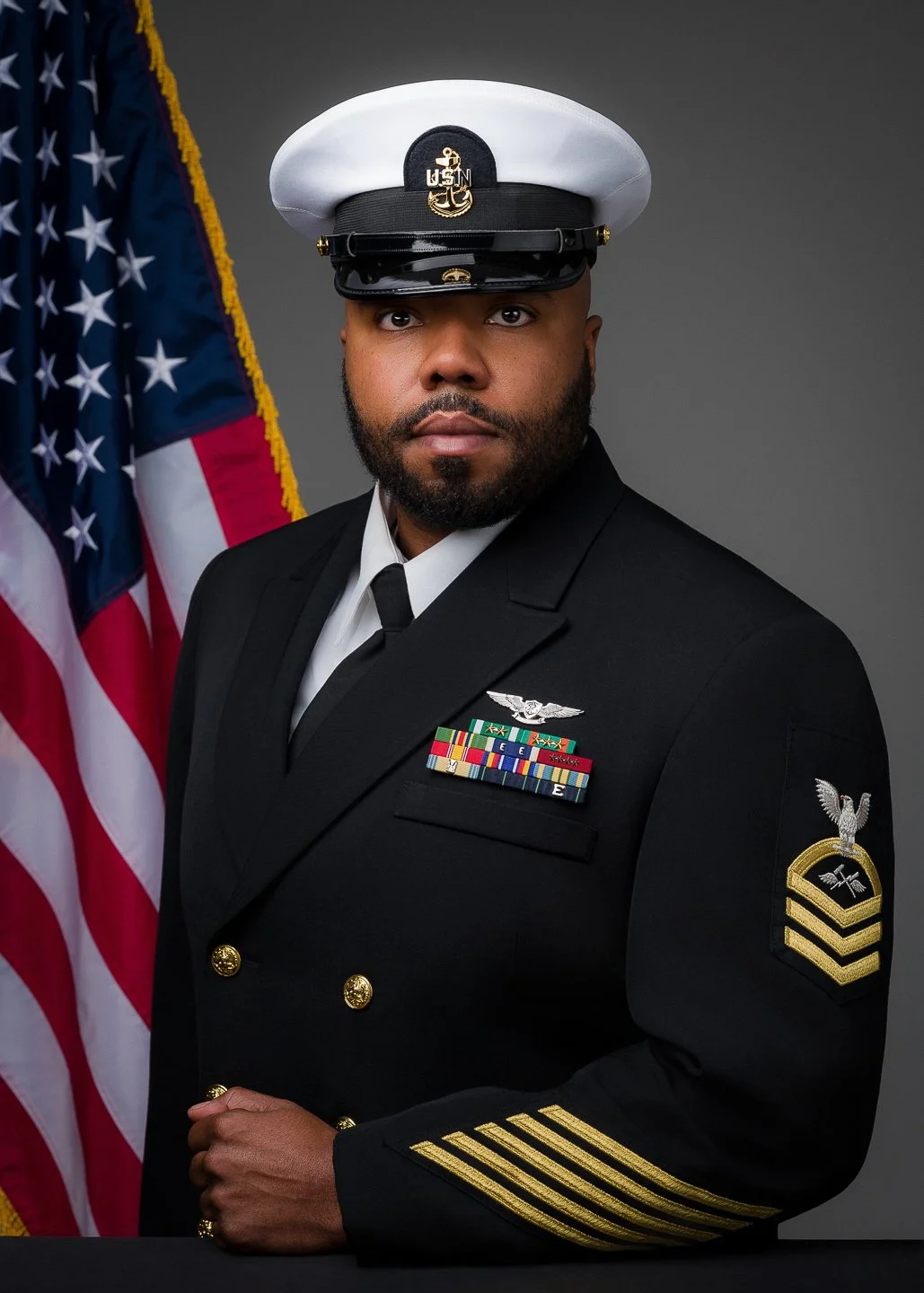 A man in a United States Navy dress uniform standing in front of an American flag. He is wearing a white sailor cap with a black brim, a dark navy coat with gold buttons, and various medals and insignia on his chest and sleeve.