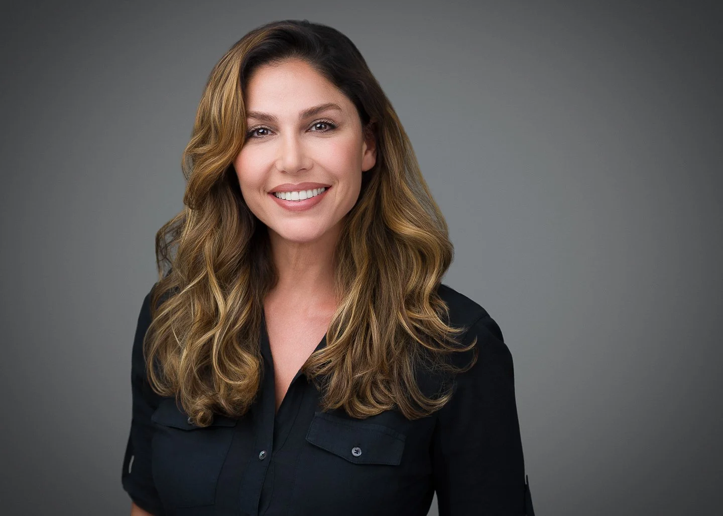 A woman with long wavy brown hair smiling, wearing a black button-up shirt against a gray background.