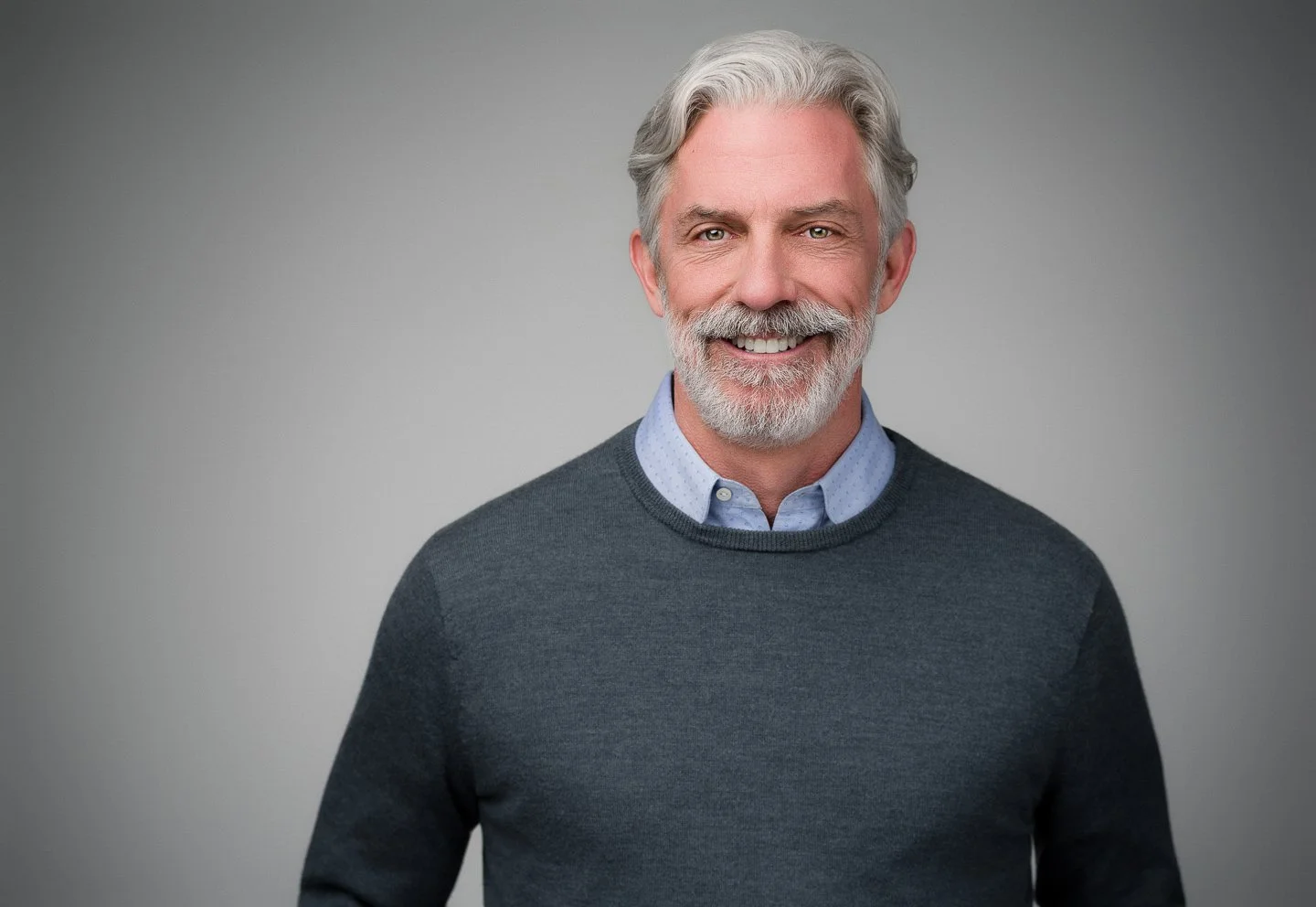 Portrait of a smiling older man with gray hair and beard, wearing a blue collared shirt and a dark gray sweater, against a plain gray background.