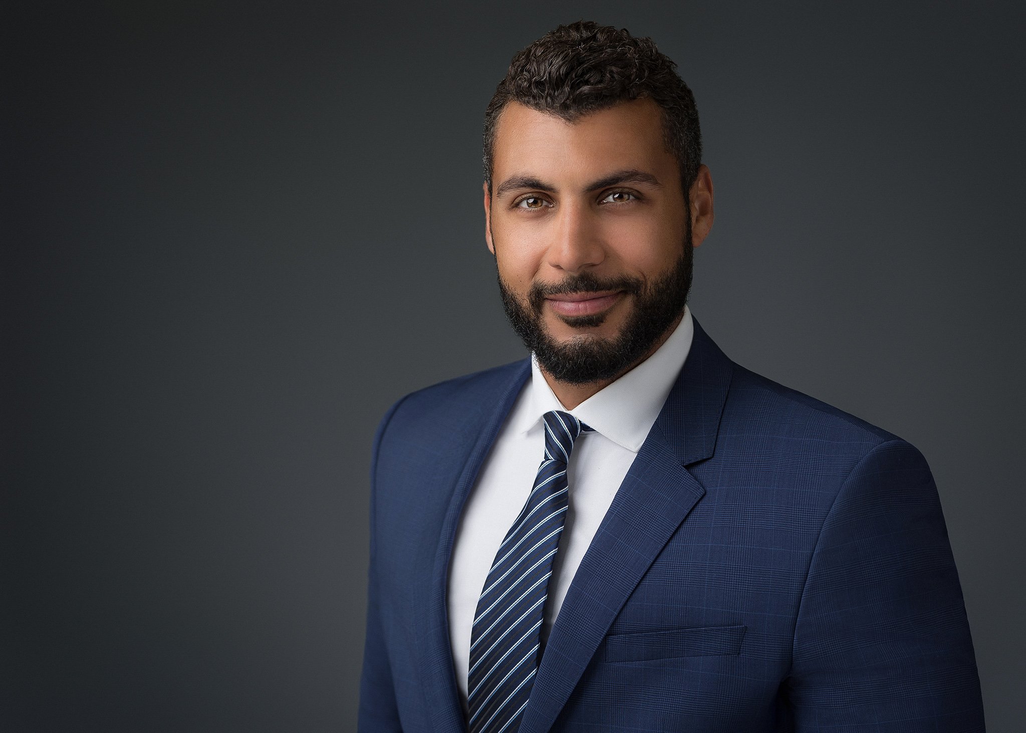 A man with dark curly hair and a beard wearing a dark blue suit, white shirt, and striped tie posing against a dark gray background.
