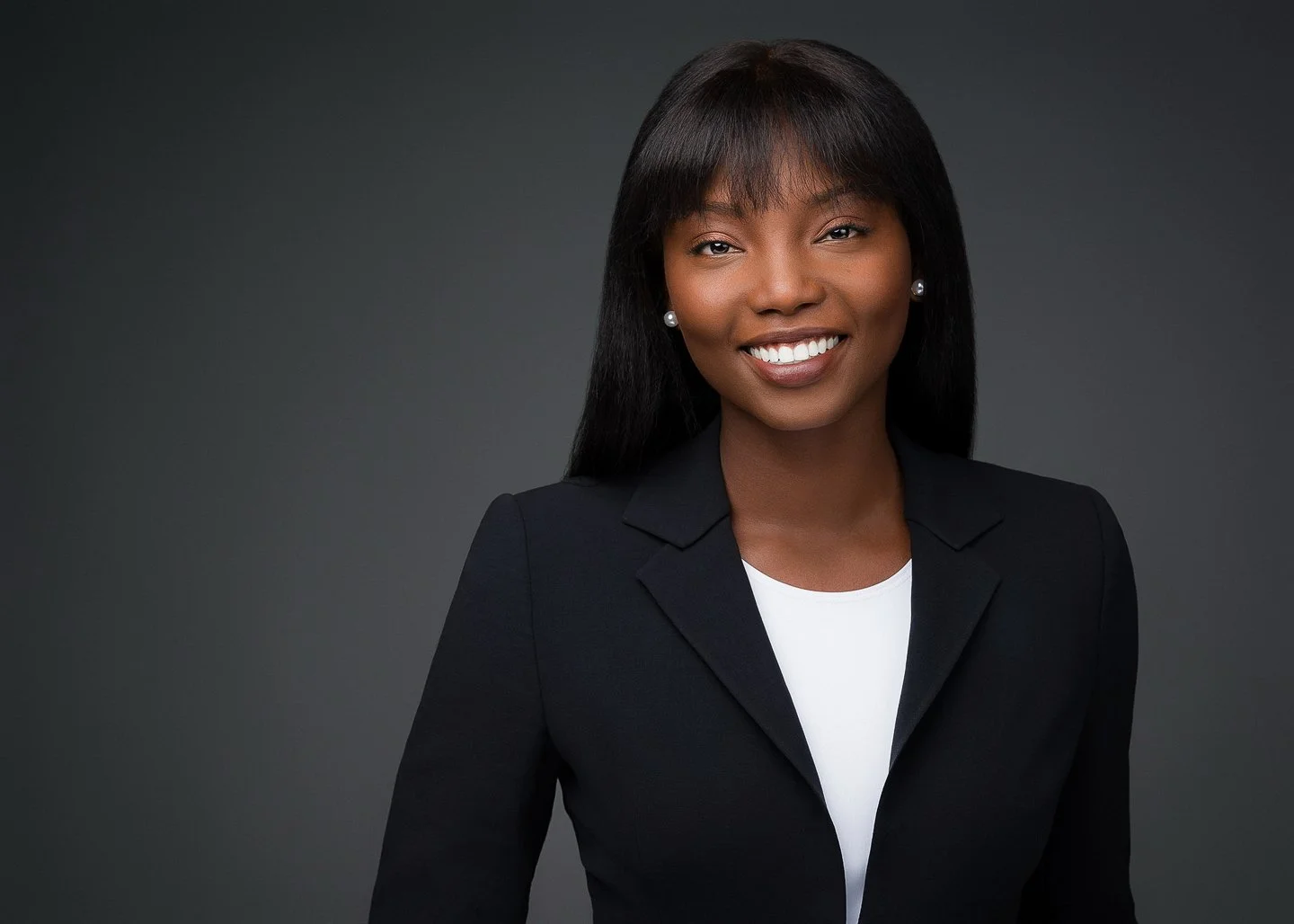 Professional woman with dark hair, smiling, wearing a black blazer over a white top, against a dark gray background.