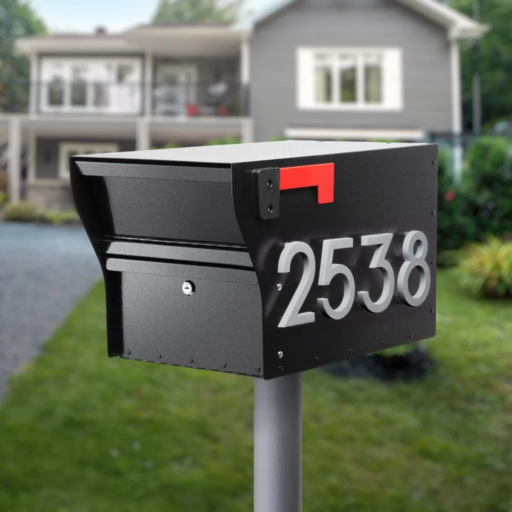 Black mailbox with house number 2538 and a red flag on a gray post in front of a house with white trim, a porch, and large windows, surrounded by greenery.