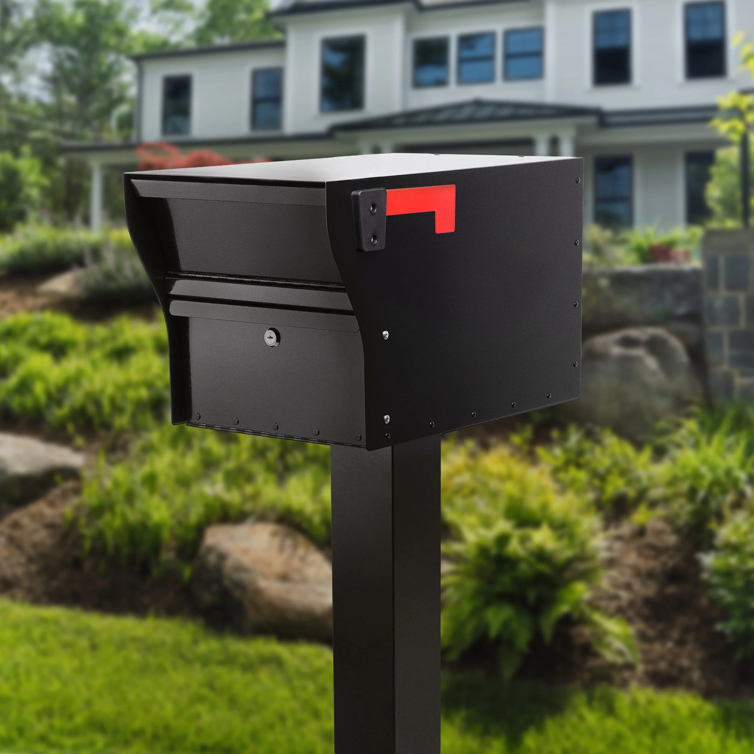 Close-up of a black mailbox with a red flag, mounted on a black post in front of a suburban house with a white exterior and multiple windows, surrounded by trees and greenery.