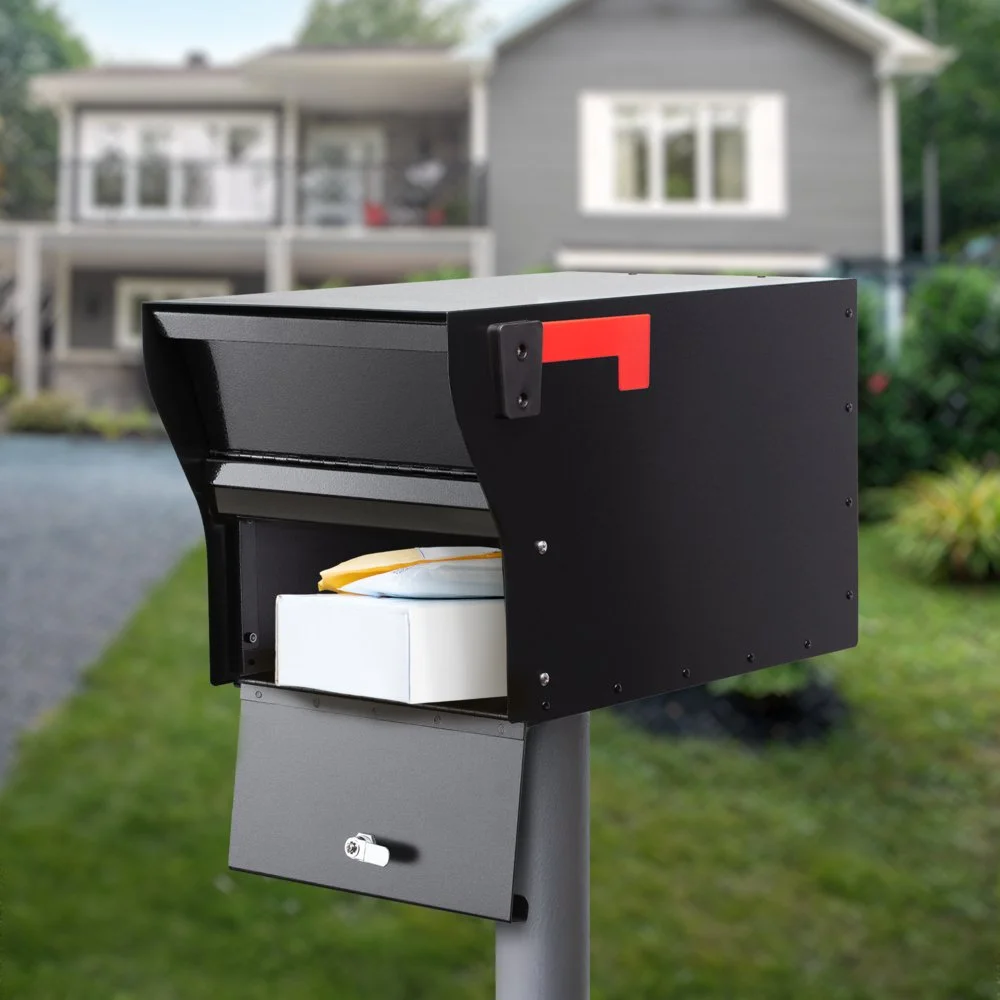 A black modern mailbox with a red flag on a gray post in front of a house with a porch and white window trim, surrounded by green grass and trees.