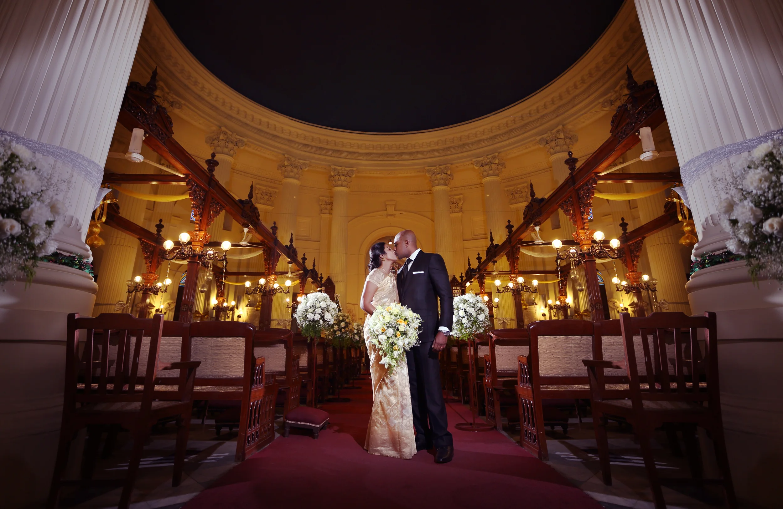 Couple in formal attire kissing inside an ornate hall with floral decorations and elegant lighting.