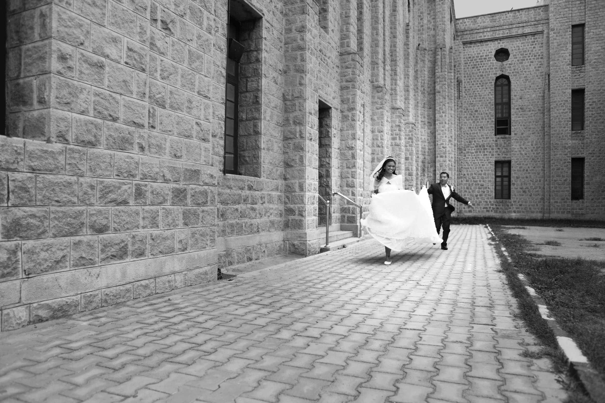 Bride and groom running on stone path next to a brick building.
