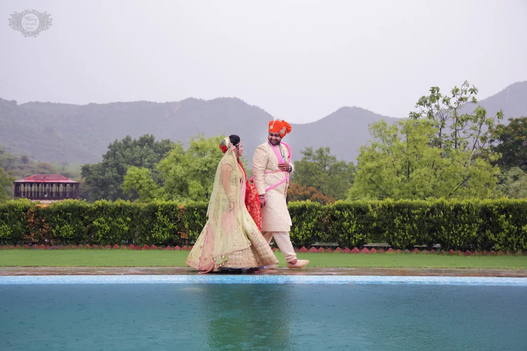 A bride and groom in traditional Indian wedding attire walking together outdoors with a scenic mountain view in the background.