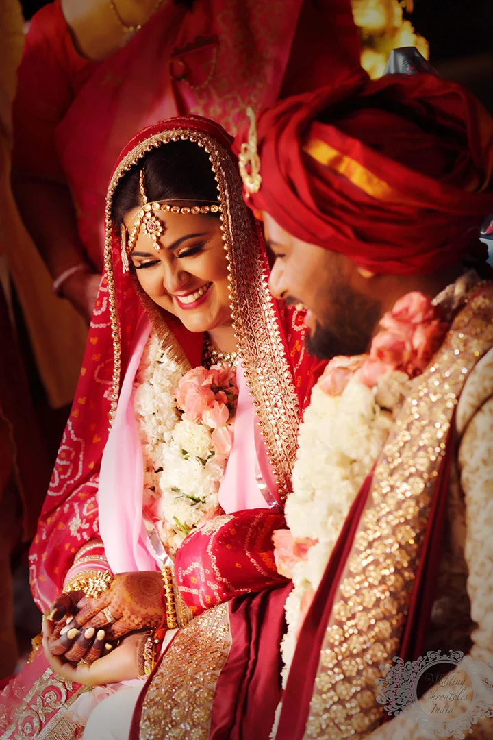 Indian bride and groom in traditional wedding attire, smiling and holding hands, adorned with floral garlands.
