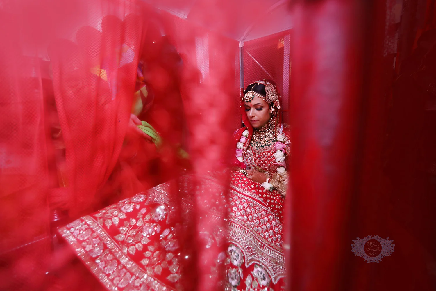 Bride in red traditional attire with jewelry, surrounded by red fabric.