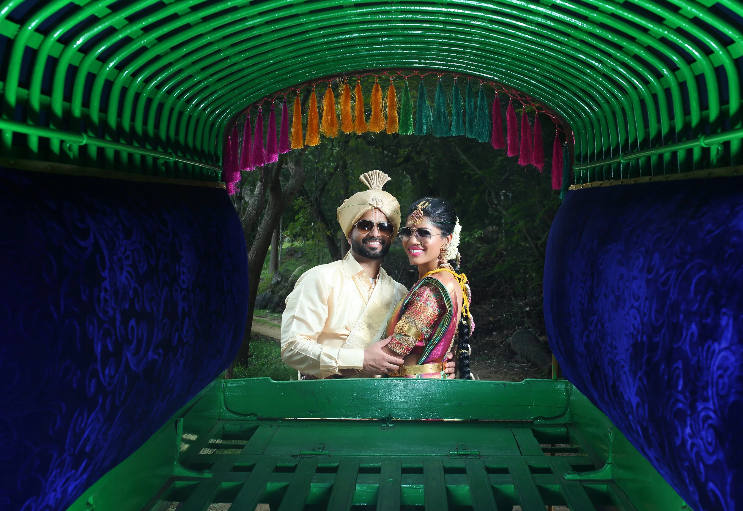 Couple in traditional attire inside a colorful cart, surrounded by green and blue fabric with tassels.