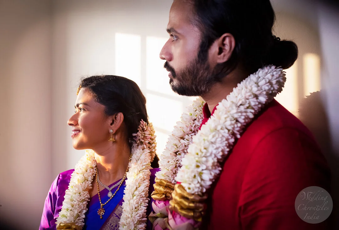 A couple wearing traditional Indian attire with floral garlands, looking towards a light source. The woman is dressed in purple and the man in red, adorned with jewelry and smiling.