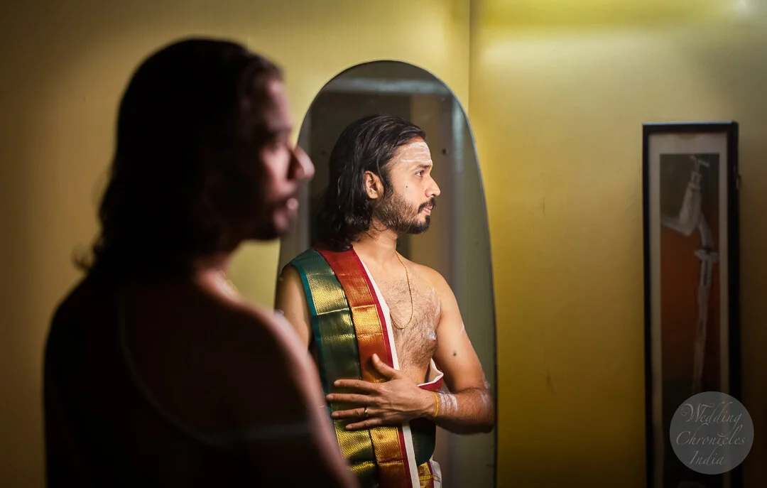 Man in traditional attire stands in front of a mirror, reflecting in it, while another man's partial reflection is seen in the foreground. The setting has a warm lighting and a framed picture is on the wall.