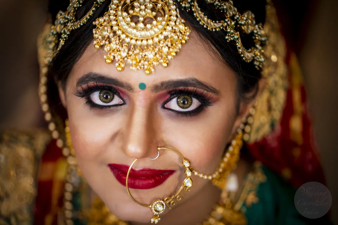 Close-up of a woman in Indian bridal attire, featuring intricate gold jewelry, a large nose ring, and traditional makeup.
