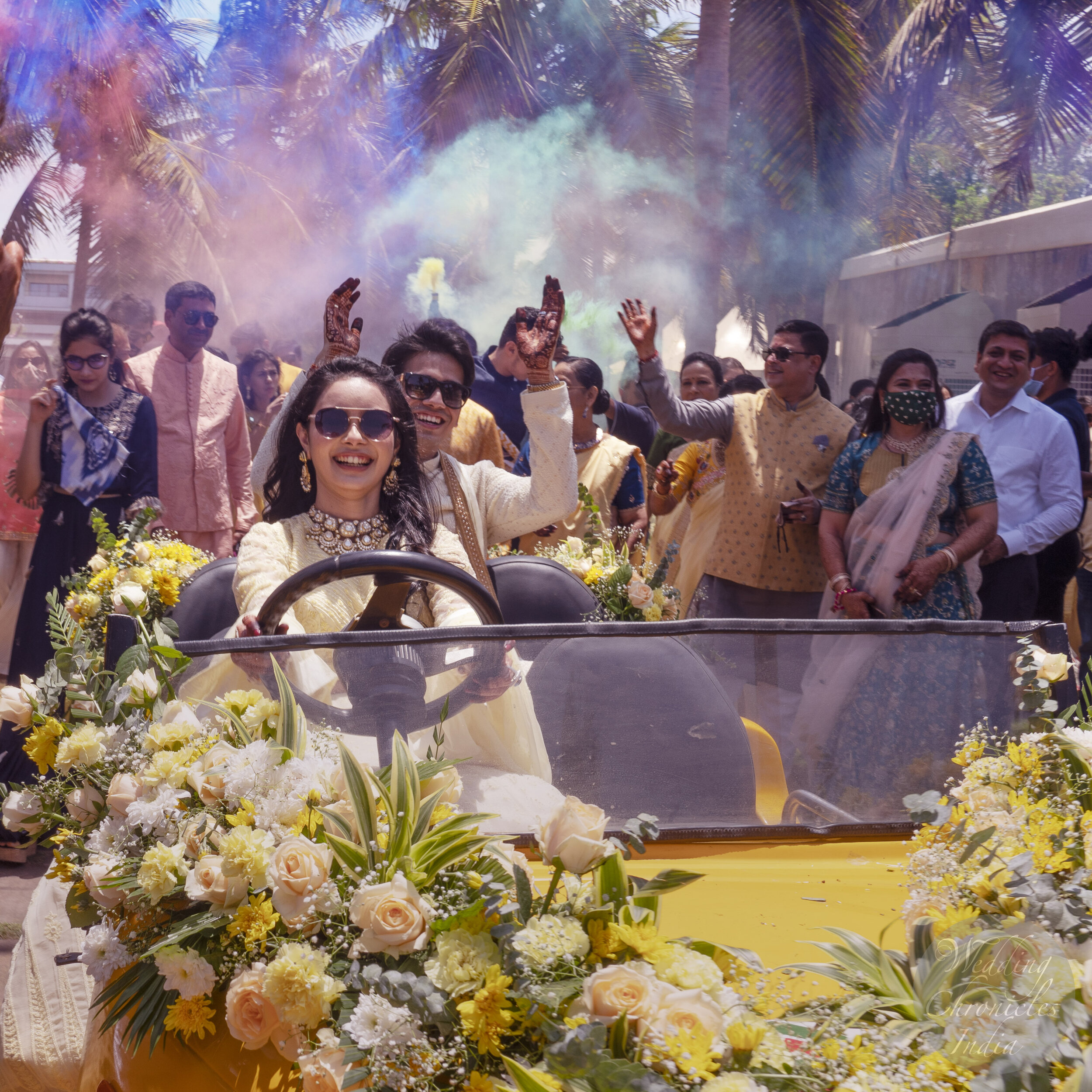 A joyful couple driving a flower-decorated cart during a festive celebration with people around them, colored smoke in the background.