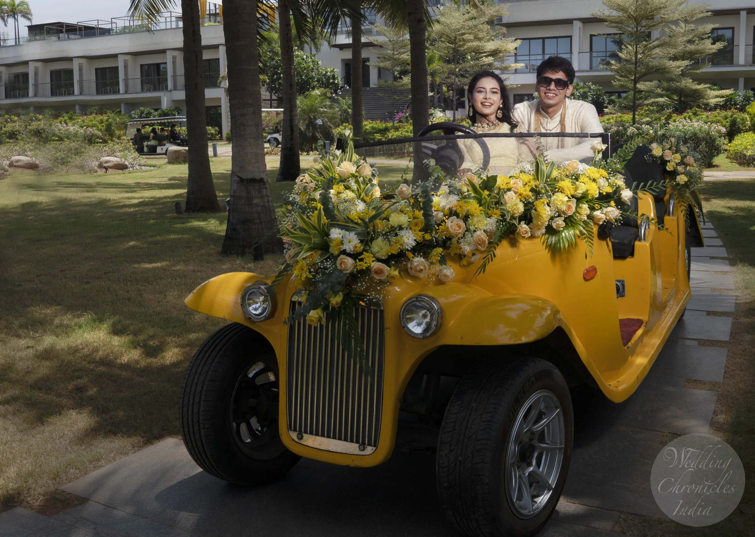Couple in vintage yellow car adorned with yellow and white flowers in a garden setting.