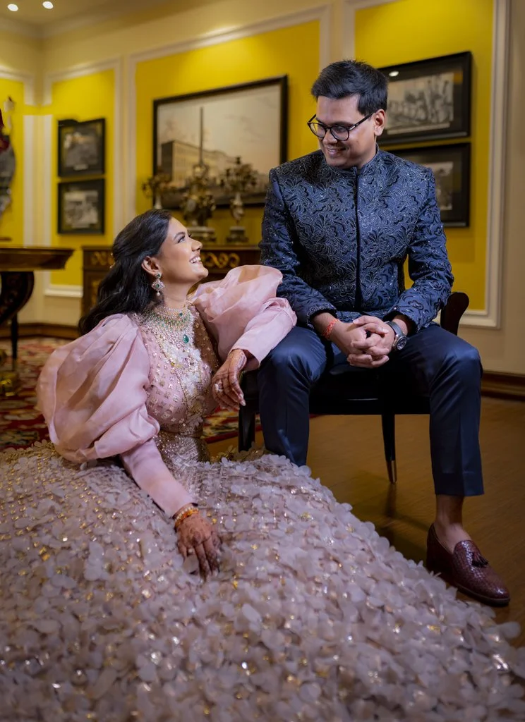 Elegant couple poses indoors; woman in elaborate pink gown sitting, smiling at man in dark blue embroidered suit seated on a chair; yellow walls and framed pictures in background.
