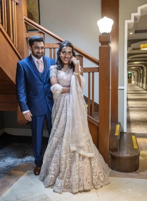 A couple portrait  standing next to a wooden staircase. The man is wearing a blue suit with a pink tie, and the woman is in an ornate cream-colored dress, smiling. The setting appears to be indoors with a lit hallway in the background.
