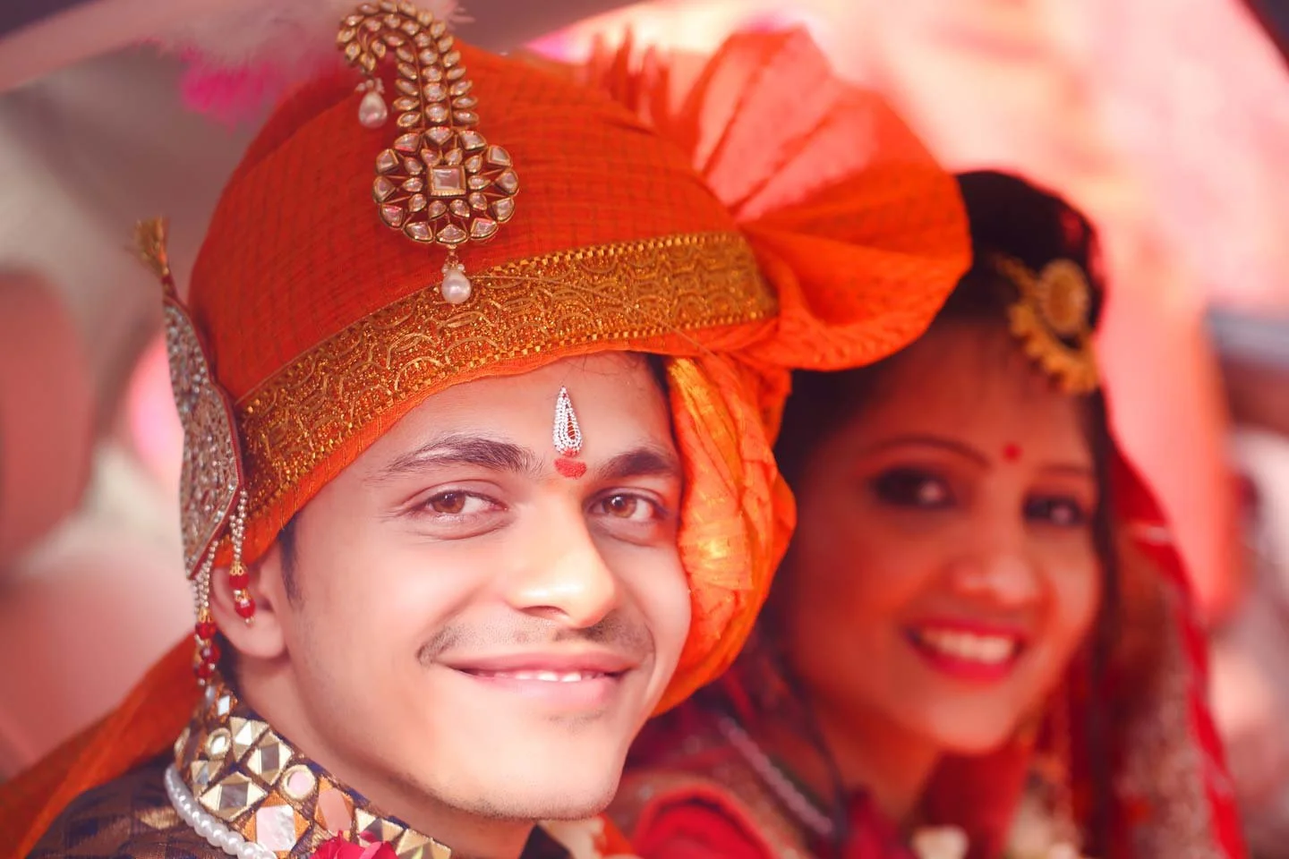 Groom and bride smile into the camera as they exit the wedding venue in their getaway car