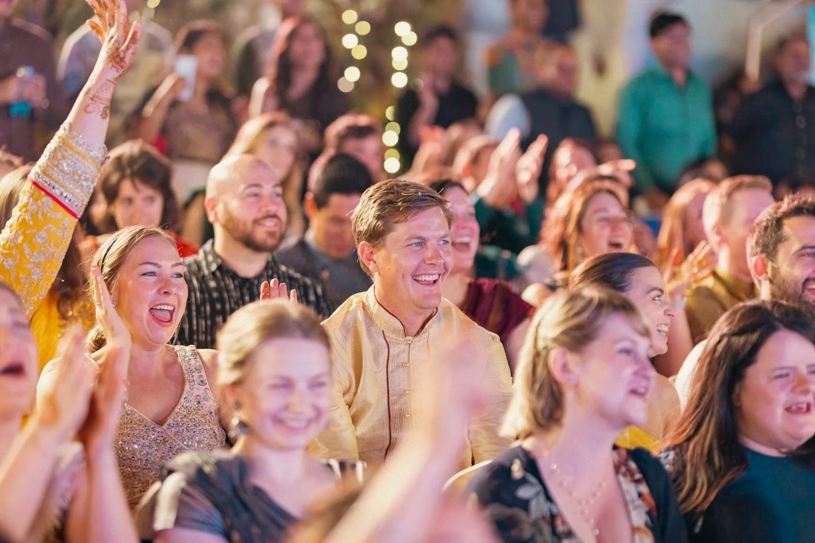 Candid photo of a guest in a crowd as he looks on and smiles during Sangeeth dance performance of the groom with his friends in Indian Wedding