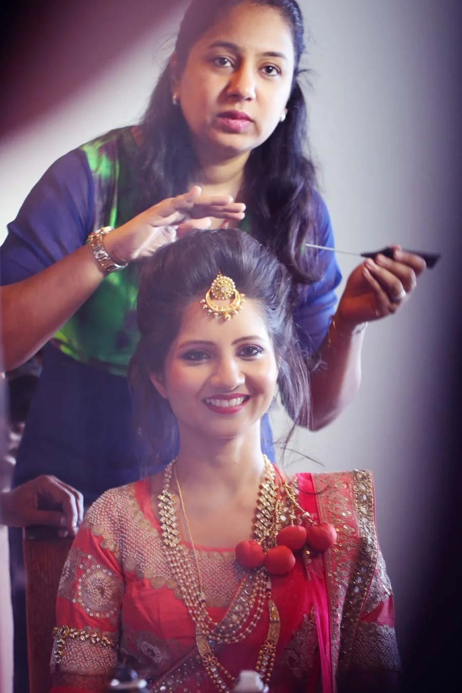 Bride looks at her reflection and smiles as make up artist does her hair