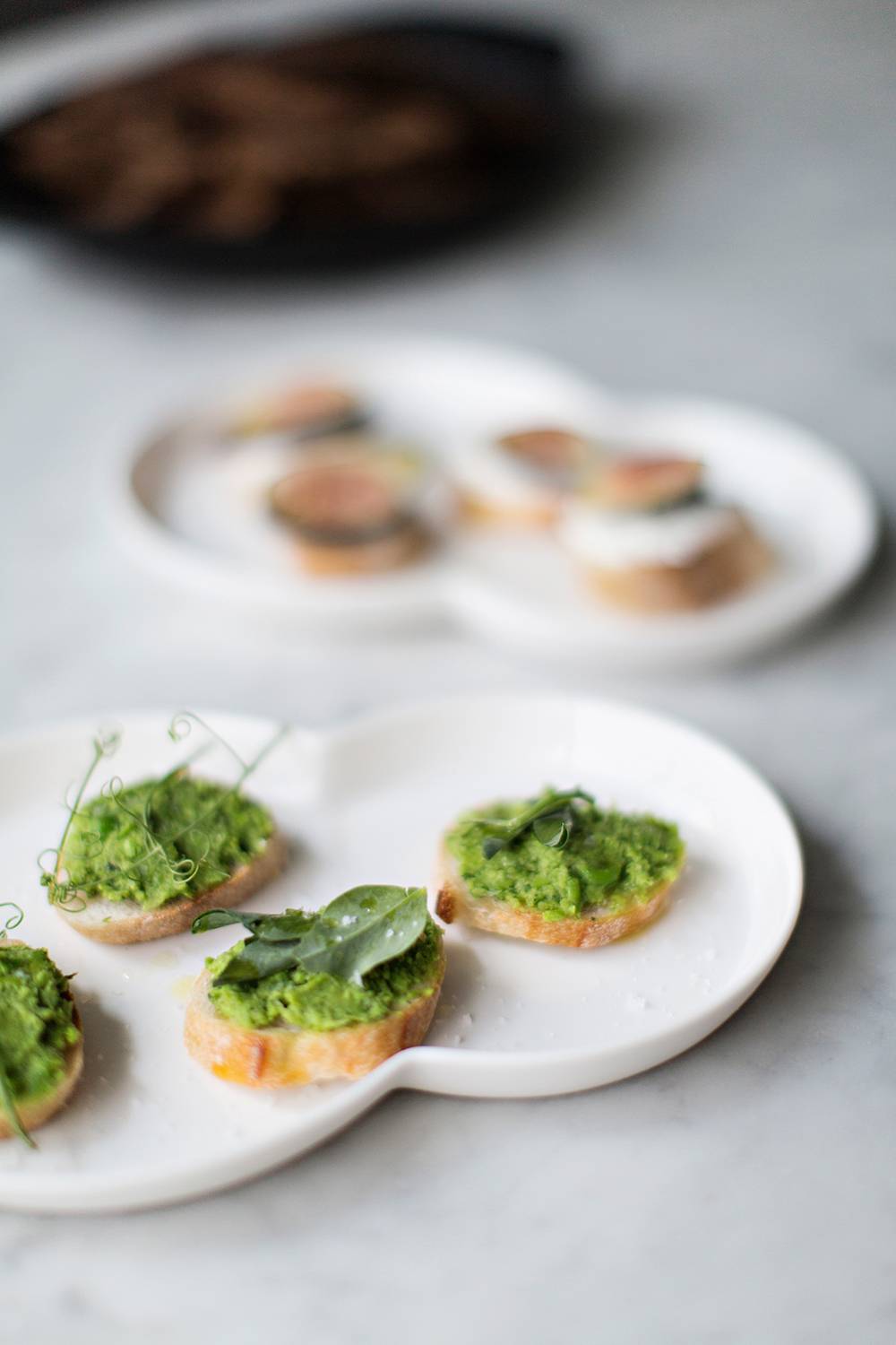 SPRING PEA CROSTINI mint + lemon 3 cups frozen peas 4 - 5 cups of water, salted 1 bunch of spring onions 1 cup fresh mint 4-5 Tablespoons of olive oil Juice of 1/2 a lemon 1 - 2 baguettes Sea salt and pepper Pea Shoots for garnish Fill a large bowl with ice water and set aside. In a large pot bring salted water to a boil. Add 3 cups of peas and boil for 2 minutes. drain peas in colander and place immediately in bowl of ice water to stop the cooking process. Set aside. Wash and dry the spring onions and cut them into thin slices. Sauté them in a hot pan with 1 Tbsp olive oil for a few minutes until they are soft but not browning. Wash the mint carefully and pick the leaves of the stems. Place the peas, spring onions, fresh mint, 3-4 spoons of olive oil and lemon juice in a blender and pulse until just blended. We prefer there peas to have a little texture so be careful not to over blend the puree. Season with salt, pepper and lemon to taste. If a smoother puree is desired then add a few teaspoons of olive oil until mixture is of desired consistency. Preheat the oven to 350 degrees. Cut the bread into slices 1 inch thick. Drizzle generously with olive oil and arrange on a baking sheet. Toast the bread for 3-5minutes. Remove from oven and set aside to cool. To serve, spread 1 tablespoon of puree over each slice of bread. Garnish with pea shoots and sprinkle with flaky sea salt and olive oil to taste. Photo + Styling | Karen Mordechai Recipe | Rikke Storm