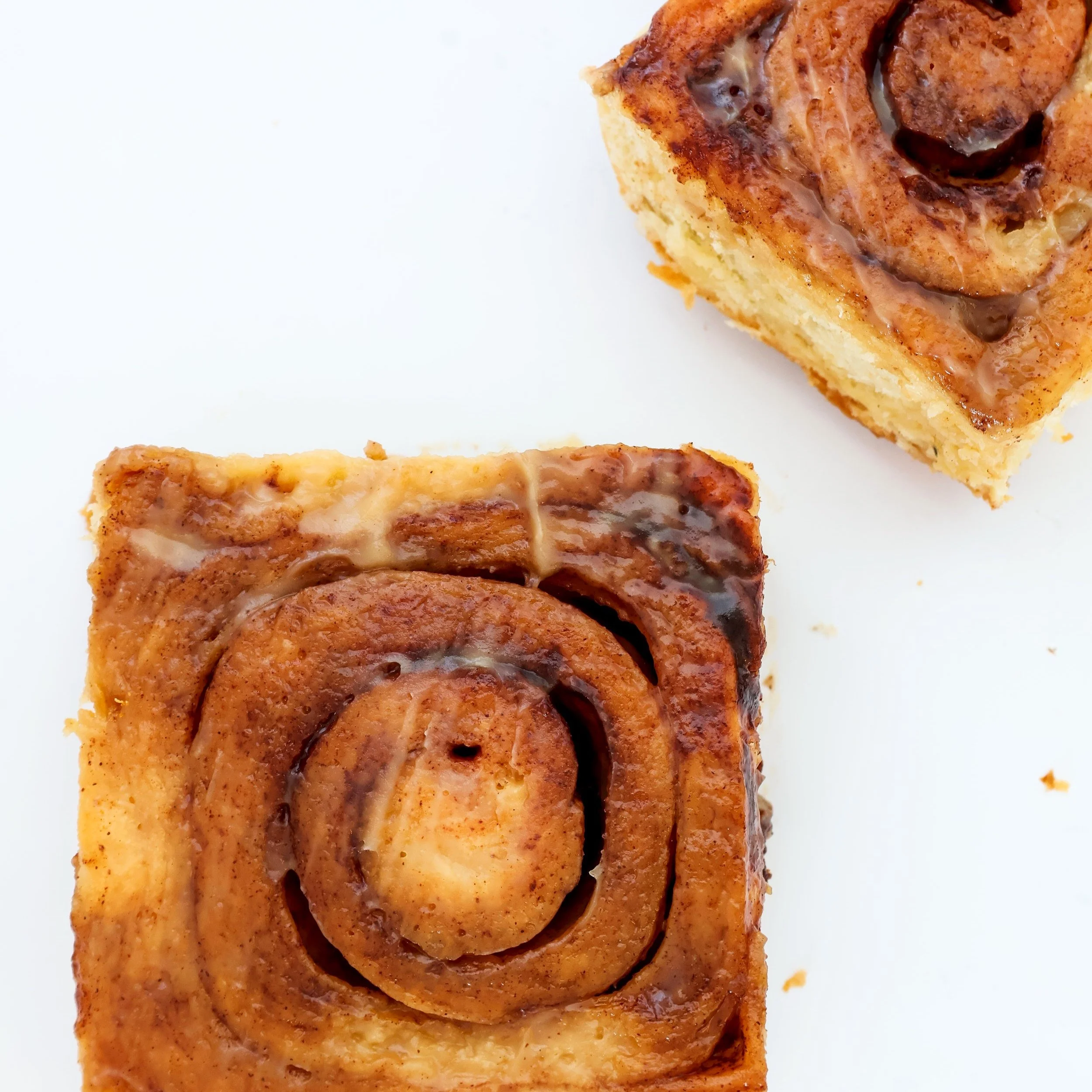 Close-up of two cinnamon rolls with icing on a white background, one near the bottom left corner and the other near the top right corner.