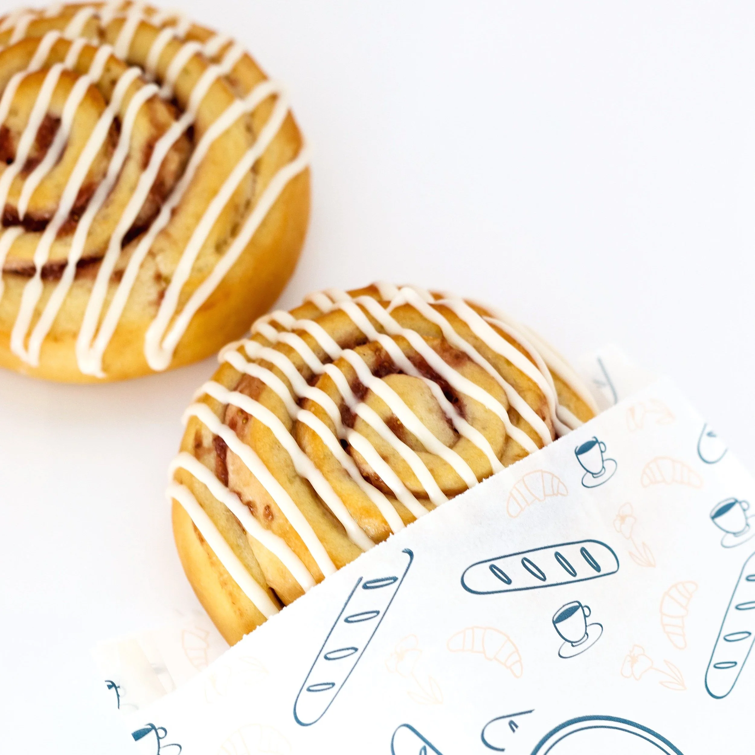 Two cinnamon rolls with icing drizzle, one partially wrapped in decorative paper, on a white surface.