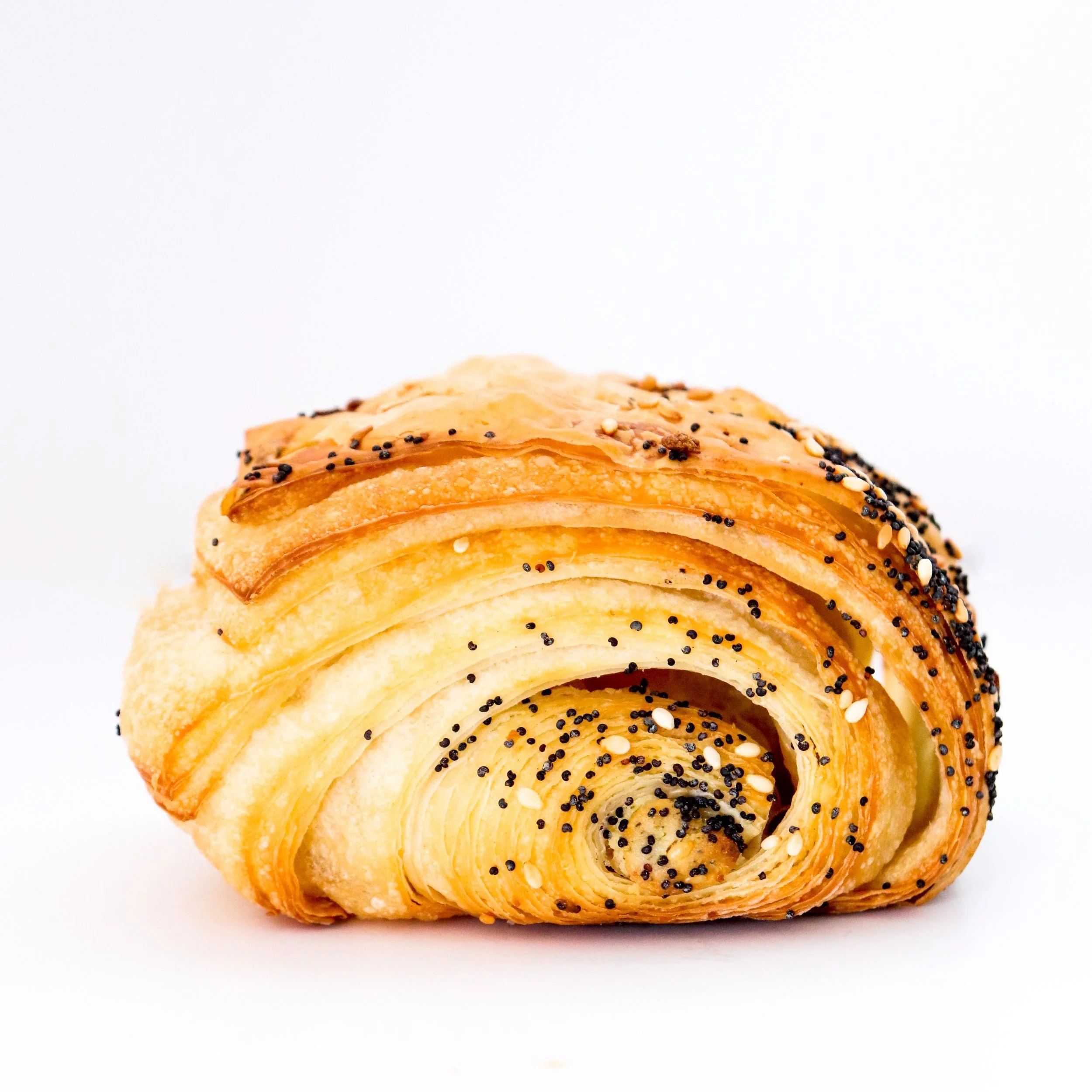 A layered, spiral-shaped pastry with a golden-brown crust, topped with black poppy seeds and white sesame seeds, on a plain white background.