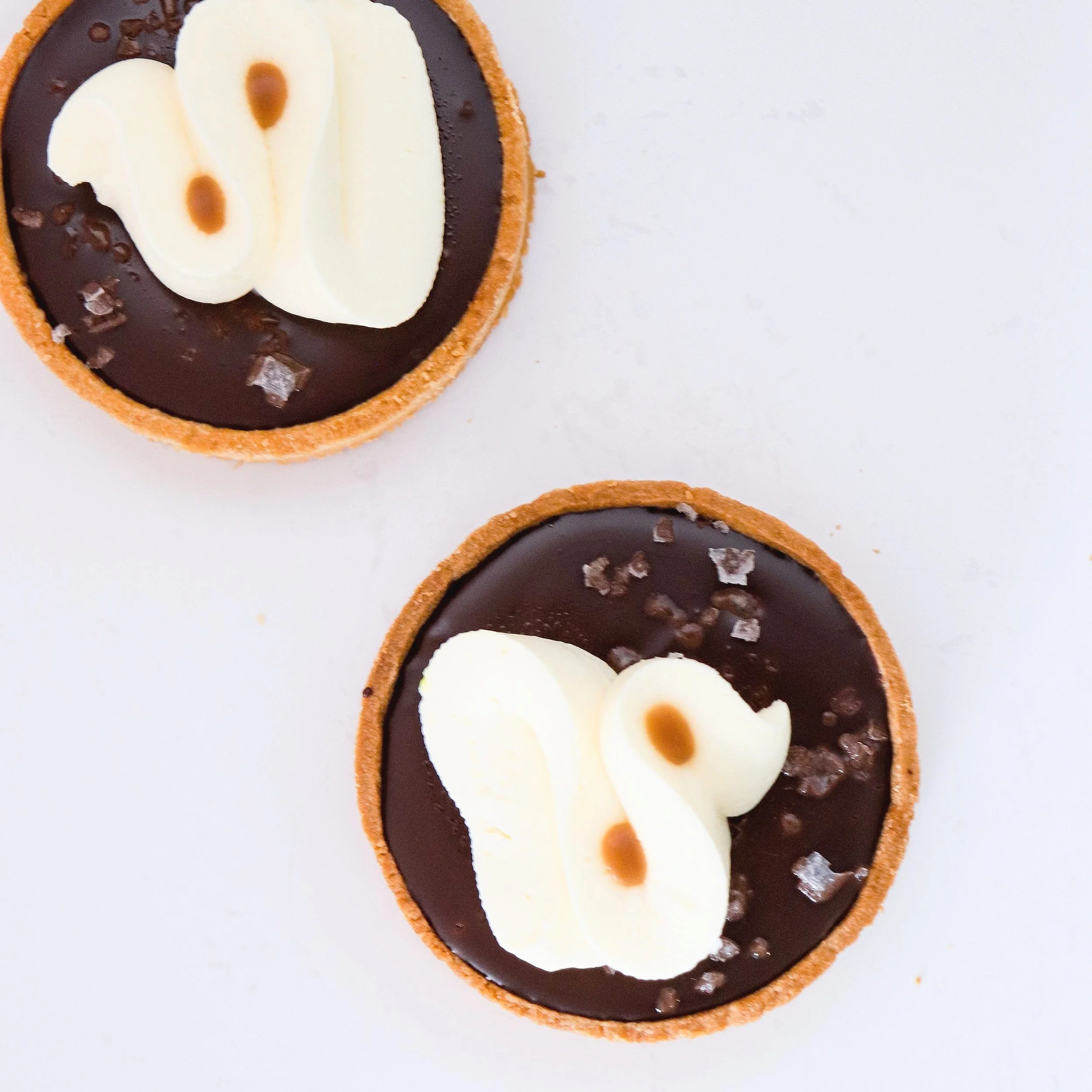 Two round chocolate tarts with white cream decoration on top, sprinkled with coarse salt, on a white surface.