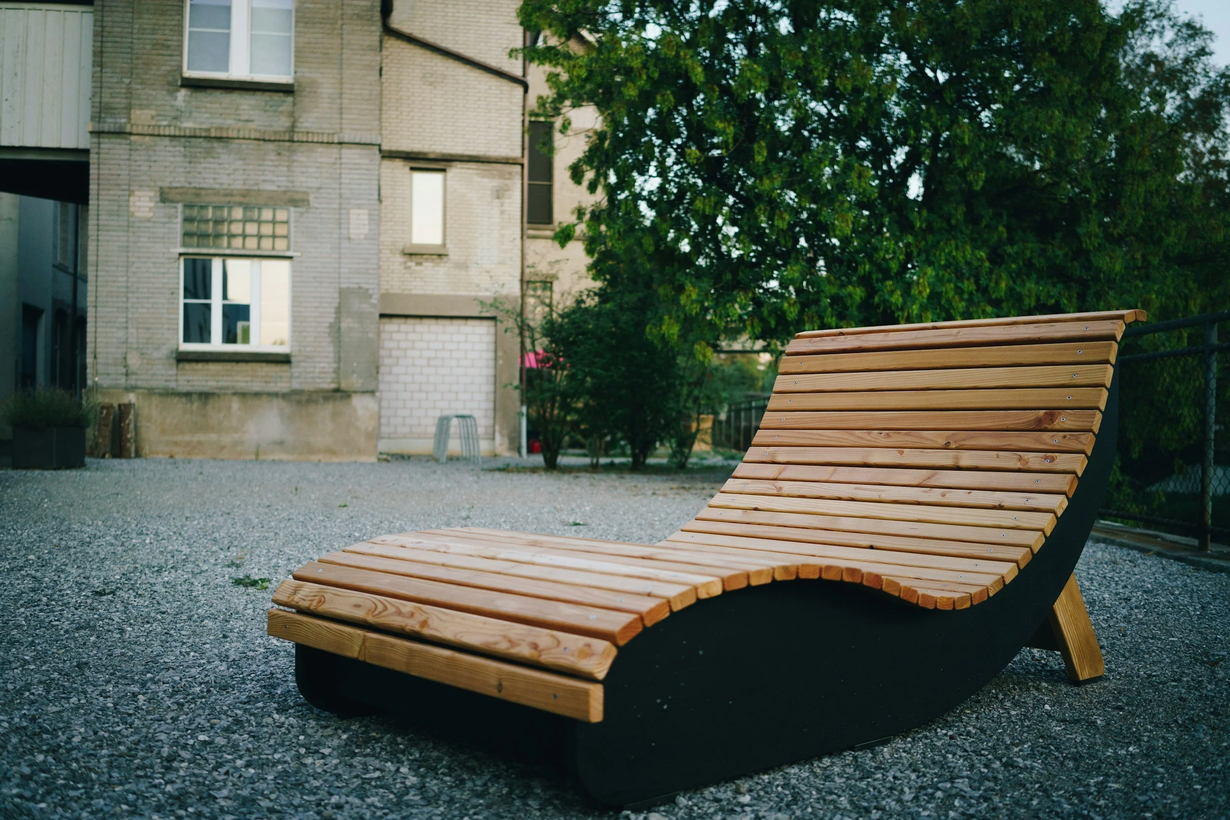 A modern outdoor wooden lounge chair with a curved design, situated on a gravel surface in a courtyard with buildings and green trees in the background.
