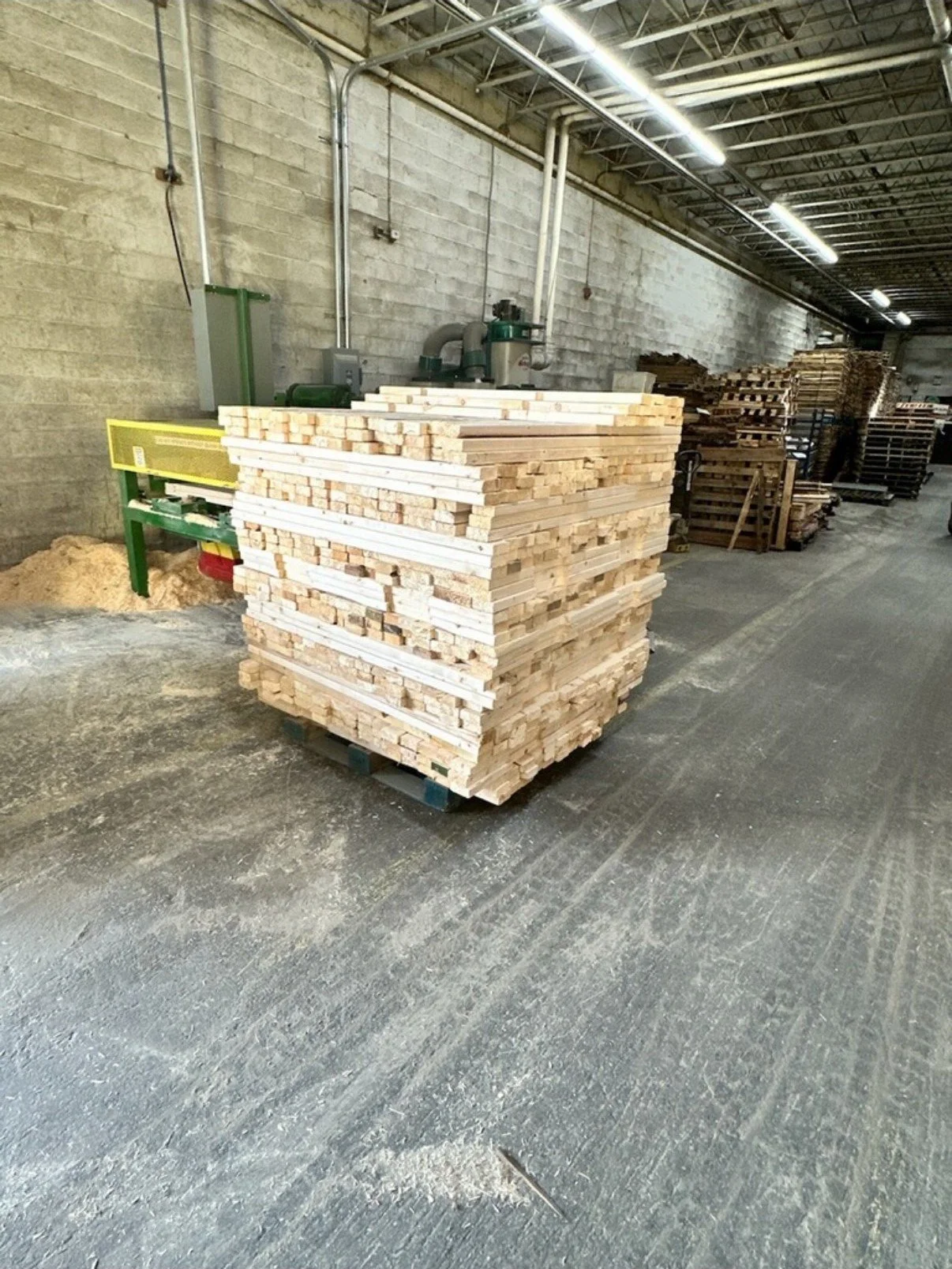 Pallet of wooden planks stored in an industrial warehouse with concrete floor and walls, and visible overhead piping and lighting.