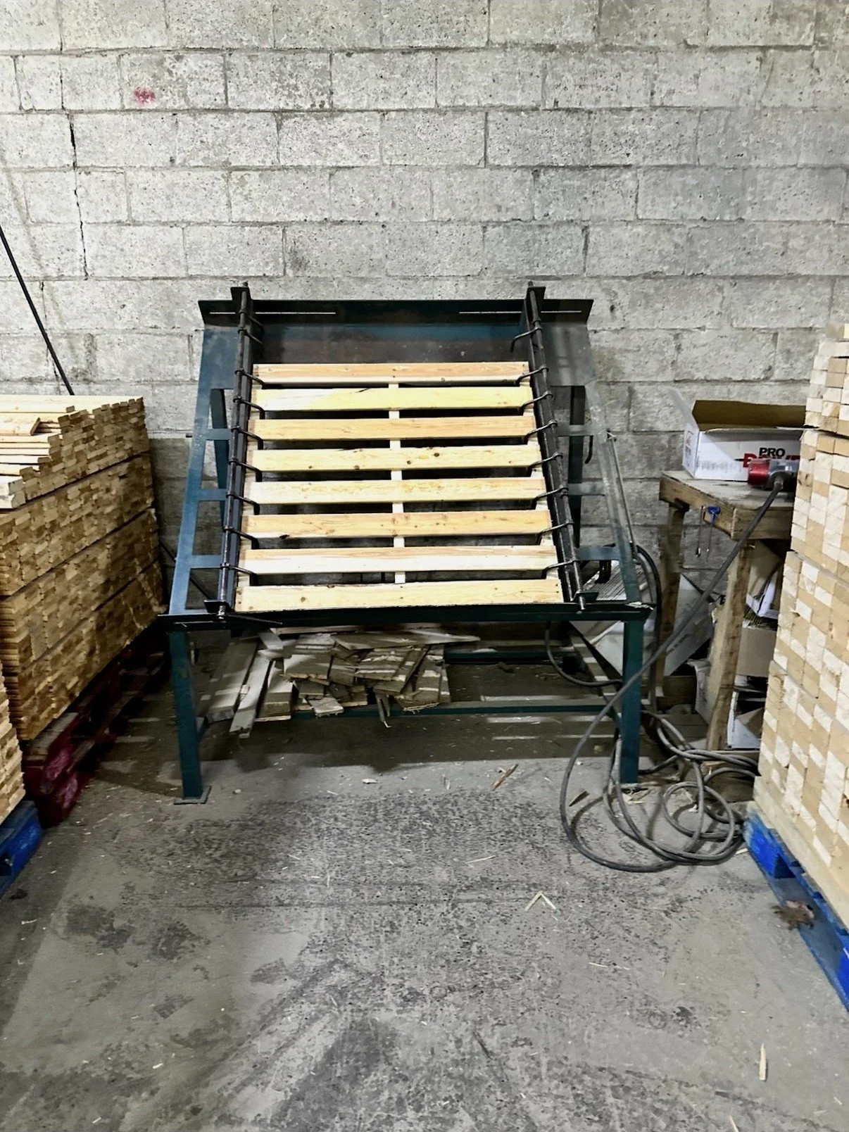 Wooden slats on a metal frame, likely a furniture or bed frame under construction in a workshop with stacks of wood and tools around.