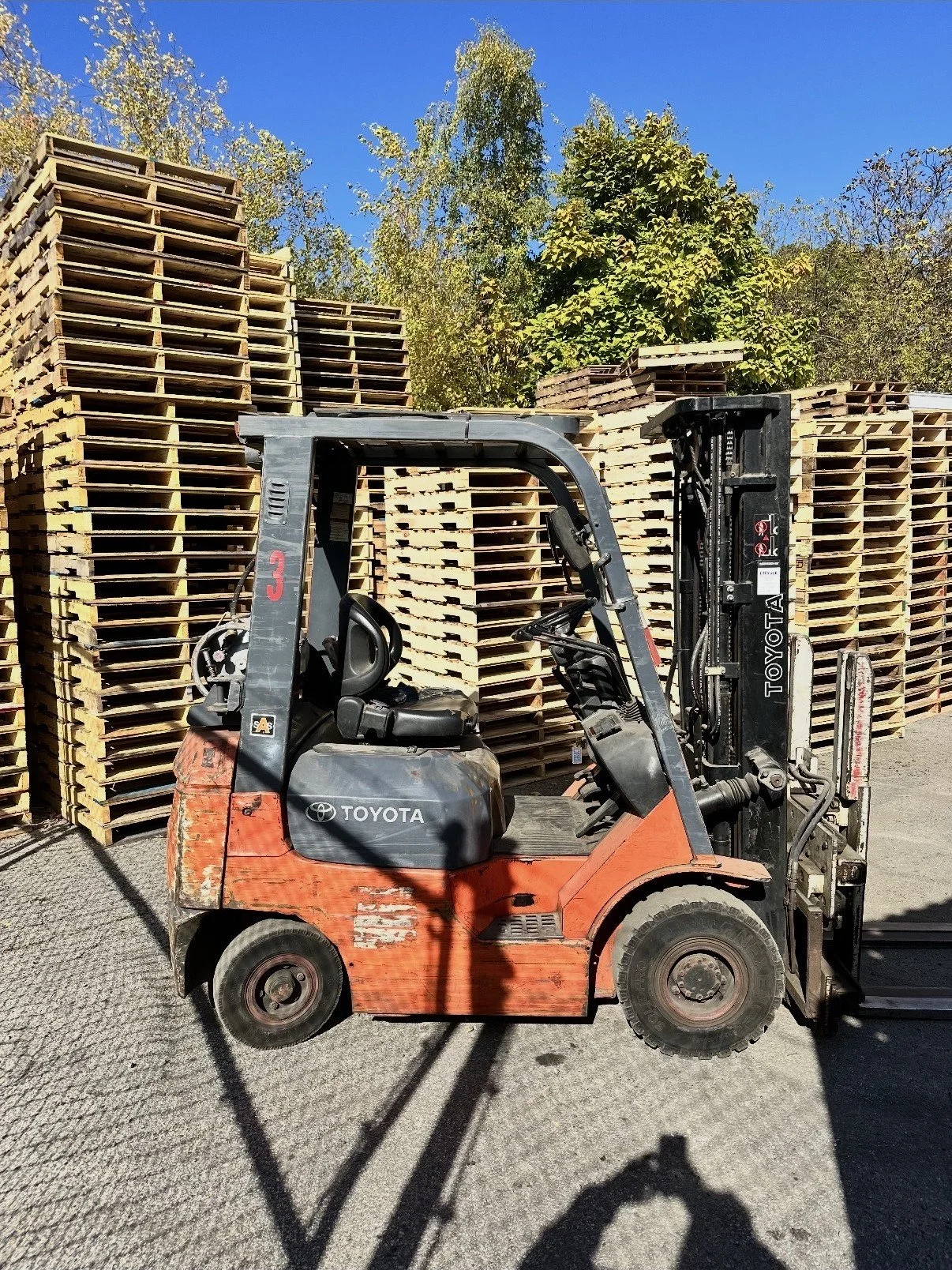 A forklift with Toyota branding parked in front of stacks of empty wooden pallets under a blue sky with trees.