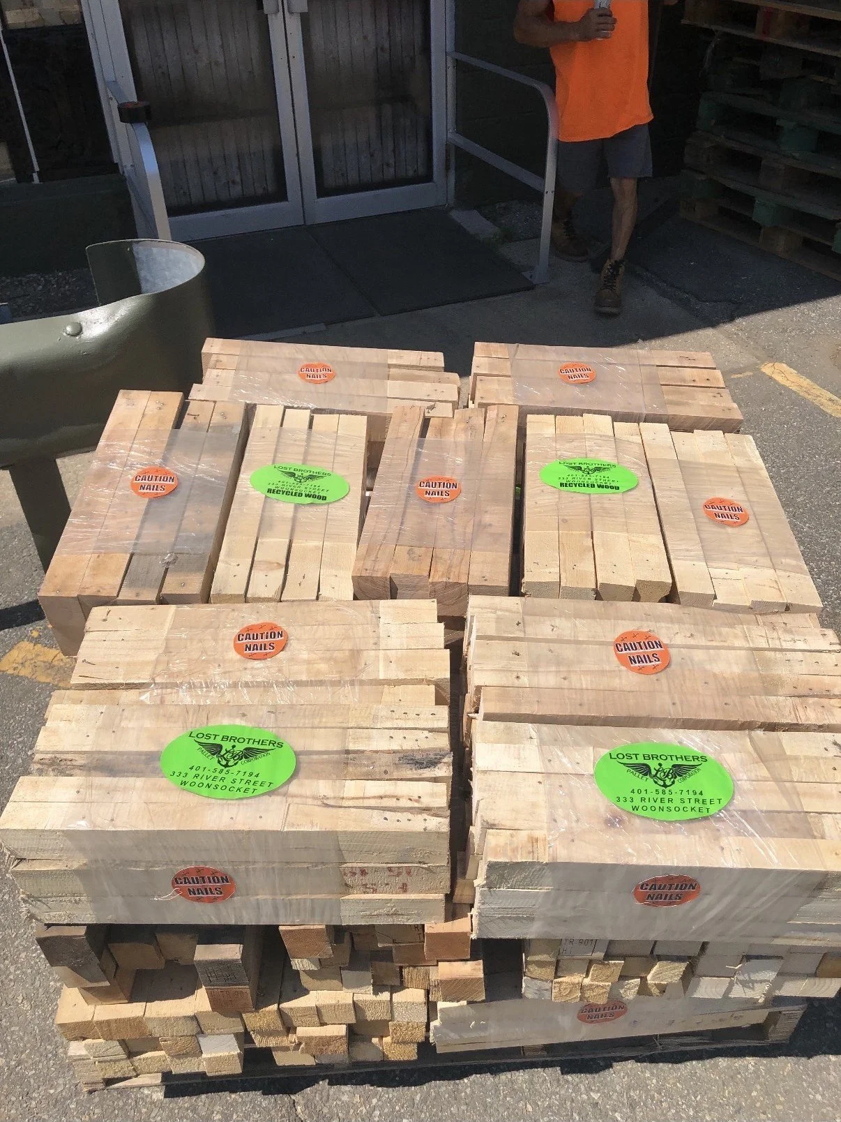 Pallets of new and recycled wood planks stacked outdoors, labeled with caution nails and recycled wood stickers, near a building entrance.