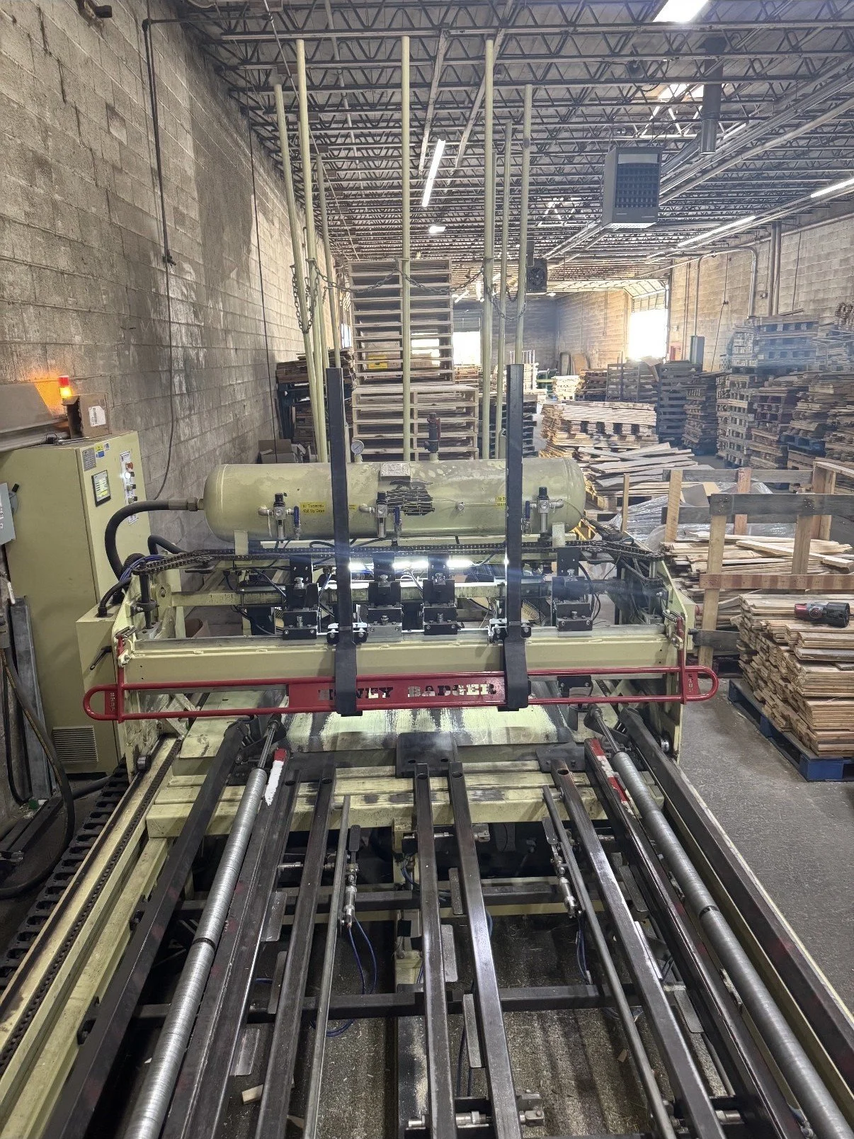 Industrial workshop with wooden pallets, stacks of wooden boards, and machinery, including a large machine in the foreground and various storage racks in the background.