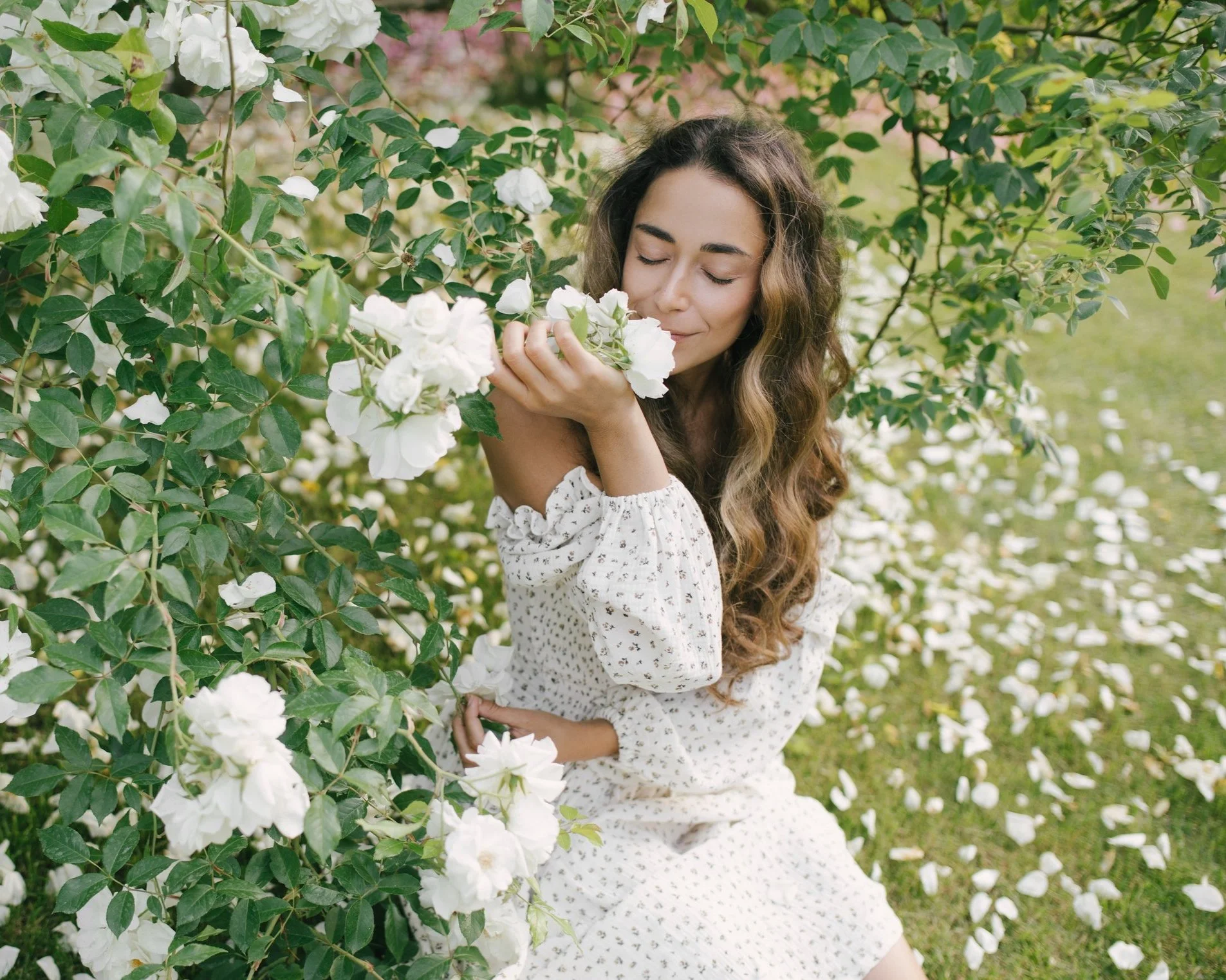 A young woman with long wavy hair smelling white flowers on a bush in a garden with green leaves and some pink flowers in the background.