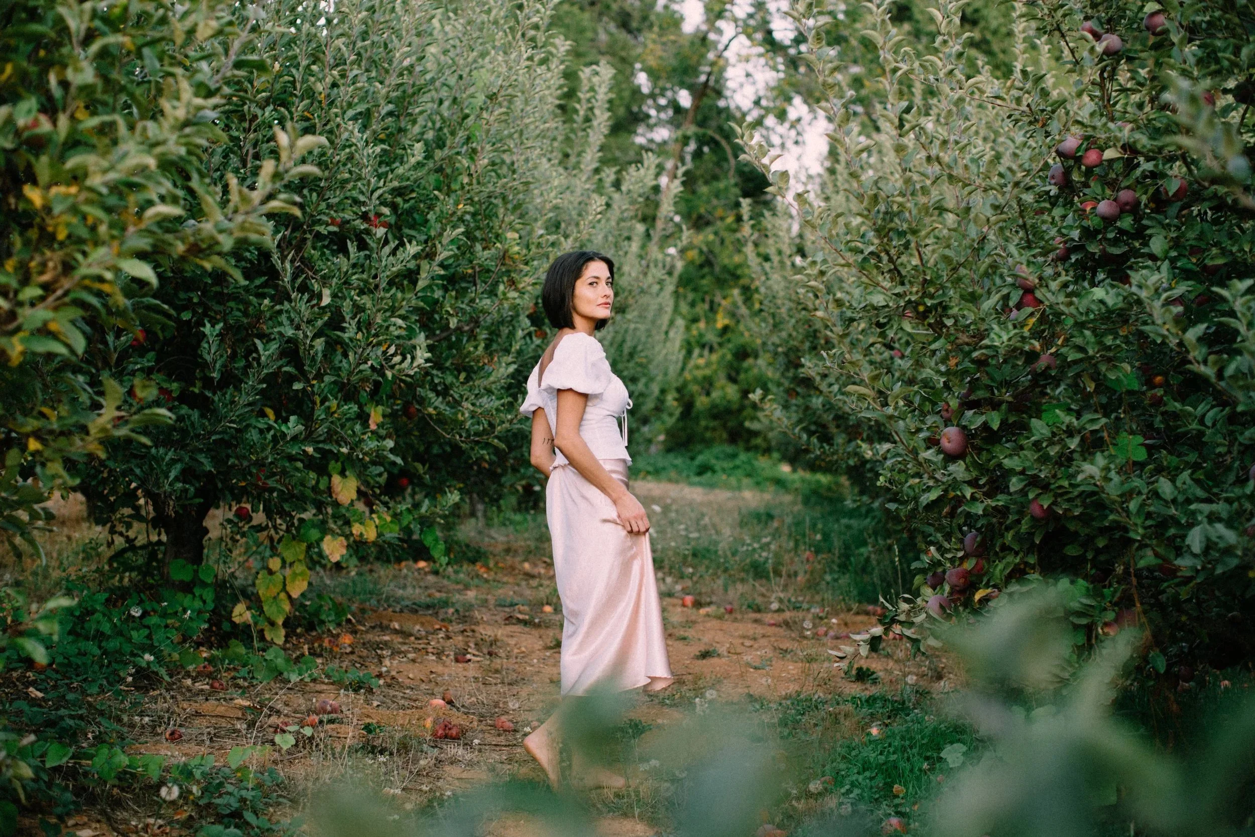 A women in a long dress standing on the earth of an apple orchard in California 