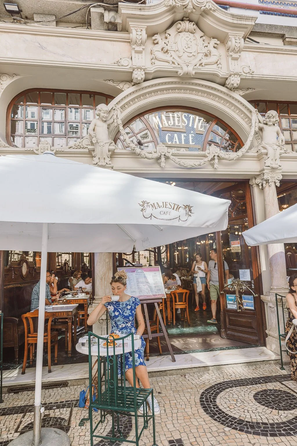 People dining outside at Majestic Cafe, with a woman in a blue floral dress sitting and sipping tea under a white umbrella, ornate architecture above the cafe entrance, and several people inside and outside.