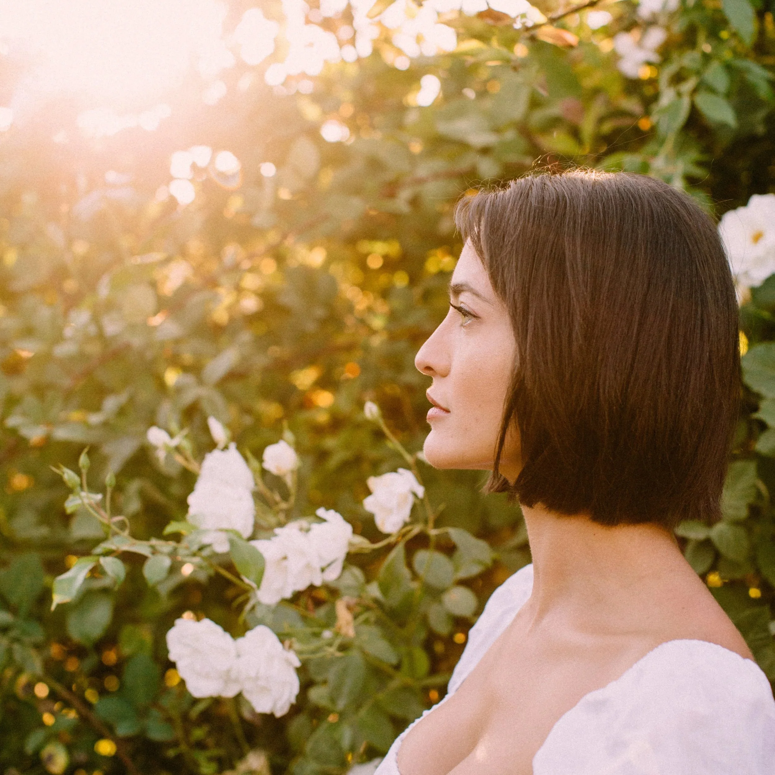 Profile of a woman with short brown hair wearing a white top, standing outdoors among green leaves and white flowers, illuminated by warm sunlight.