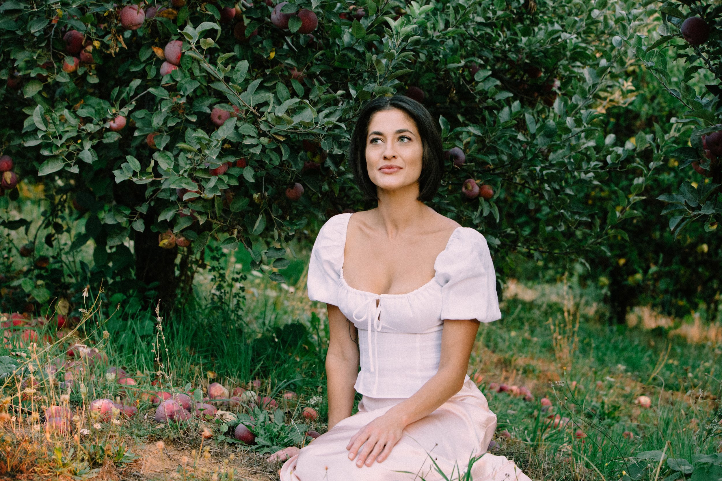 A woman with shoulder-length dark hair and light skin sits on the ground in a garden surrounded by apple trees and fallen apples. She is wearing a white, puff-sleeved top and light-colored pants, looking to her left with a serene expression.
