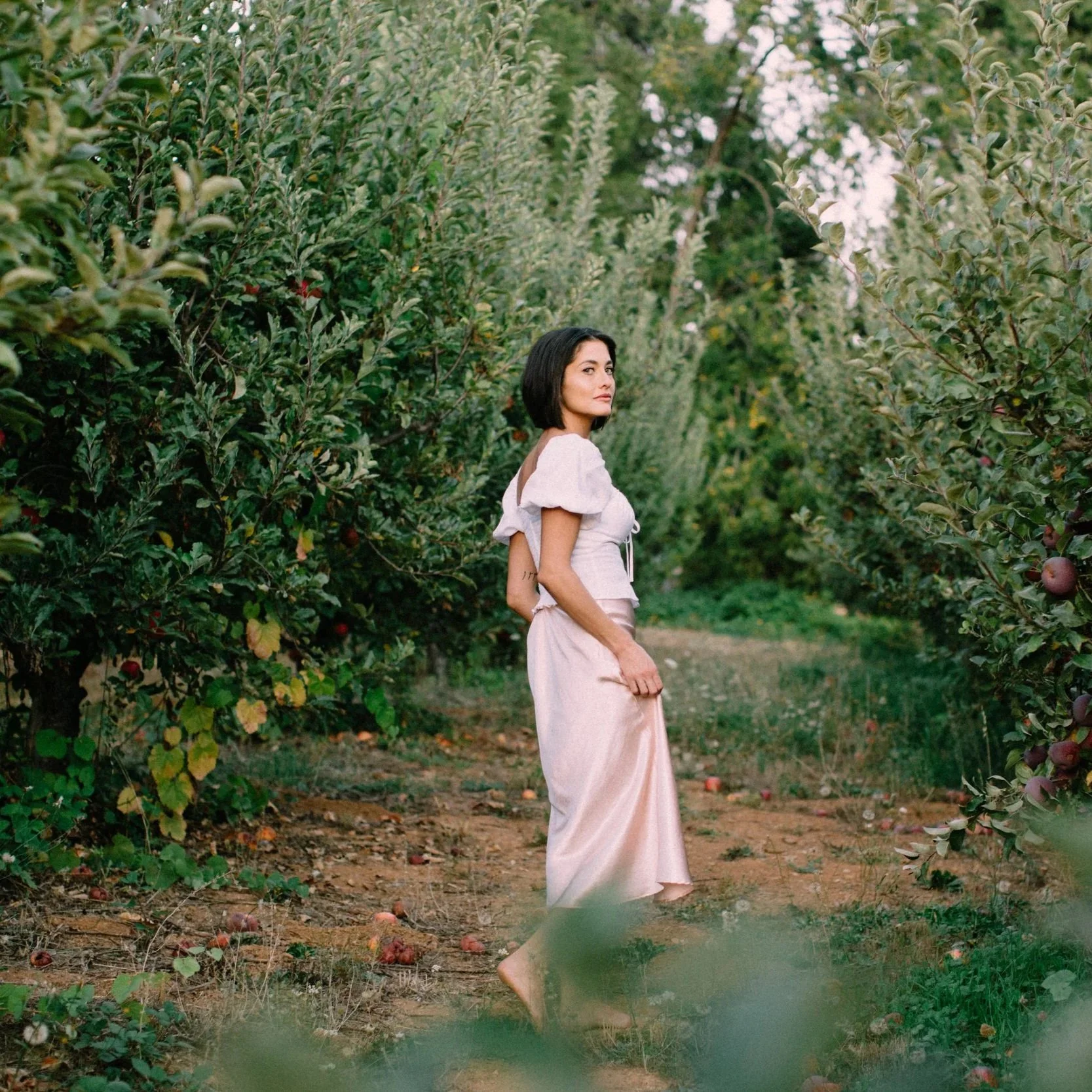 A woman with shoulder-length dark hair wearing a white top and a long pink skirt standing in an apple orchard surrounded by apple trees. She looks to the side with a calm expression, and fallen apples are on the ground.