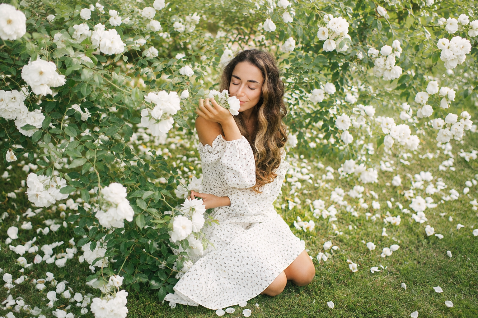 A woman with long, curly hair and a white dress kneeling beside a lush bush of white flowers, smelling one of the blossoms with her eyes closed.