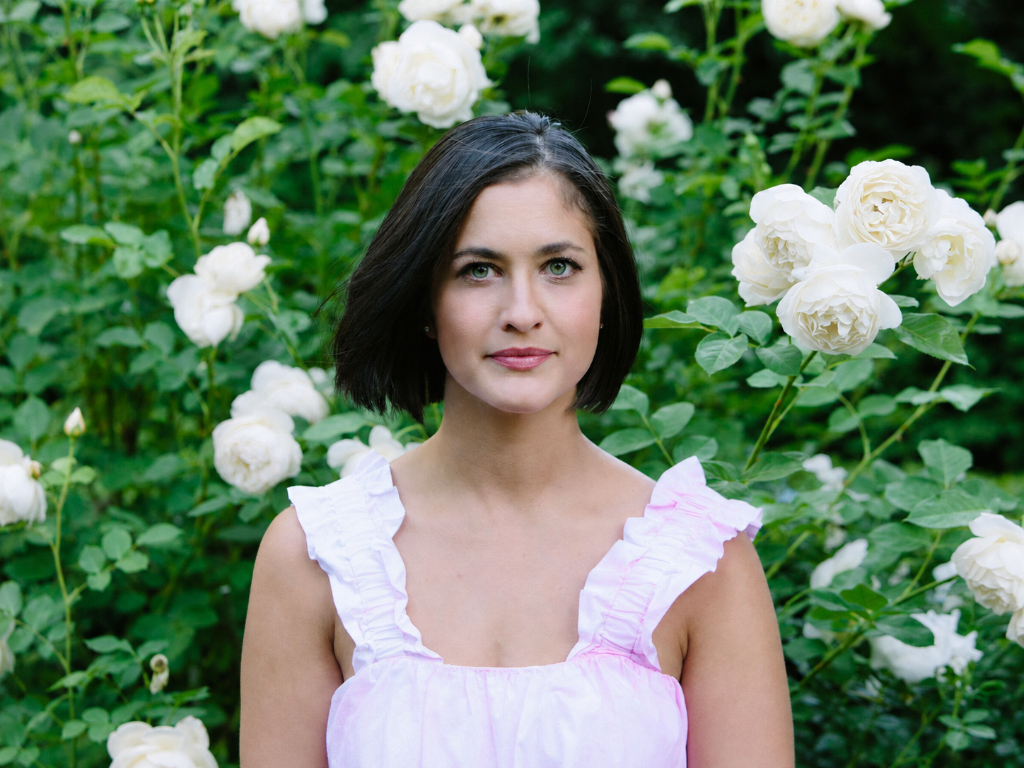 A woman with short black hair and blue eyes standing in front of a bush of white roses, wearing a pink ruffled sleeveless top.