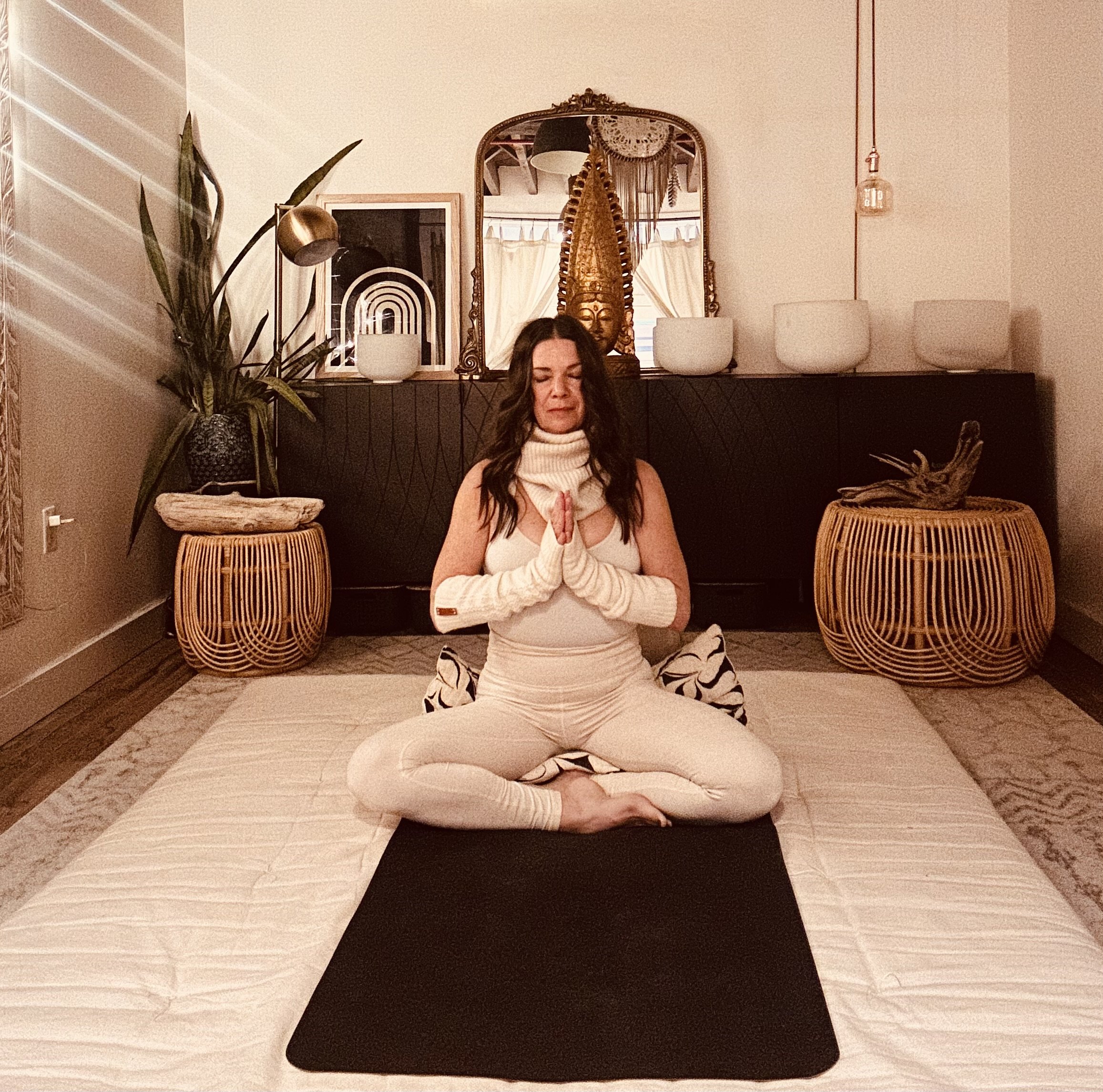 A woman practicing yoga in a meditation pose on a black mat, sitting cross-legged with hands pressed together in front of her chest, in a cozy, decor-filled room with plants, artwork, and a mirror.